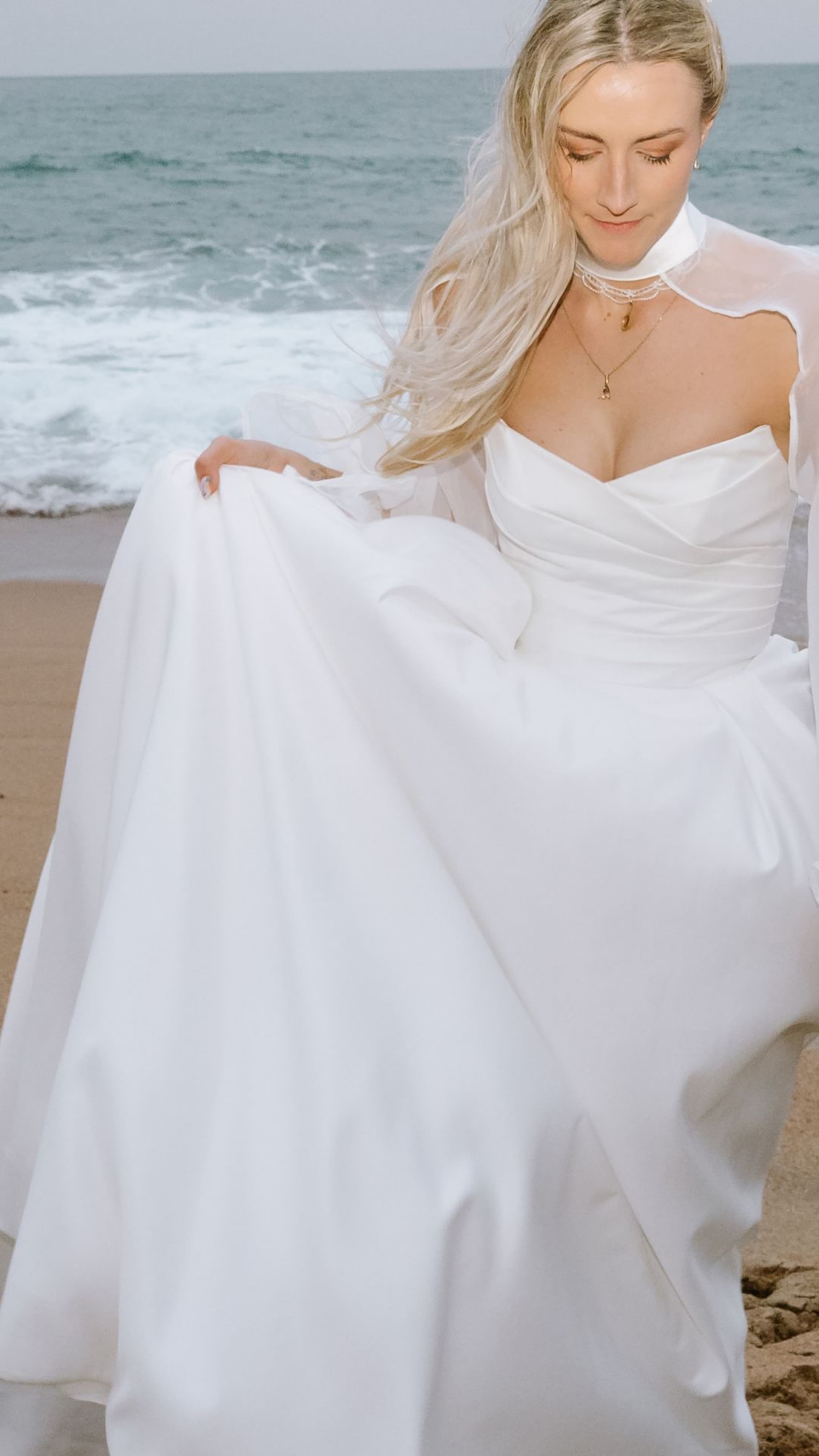 A blonde bride in a white wedding gown walks along a sandy beach near ocean waves