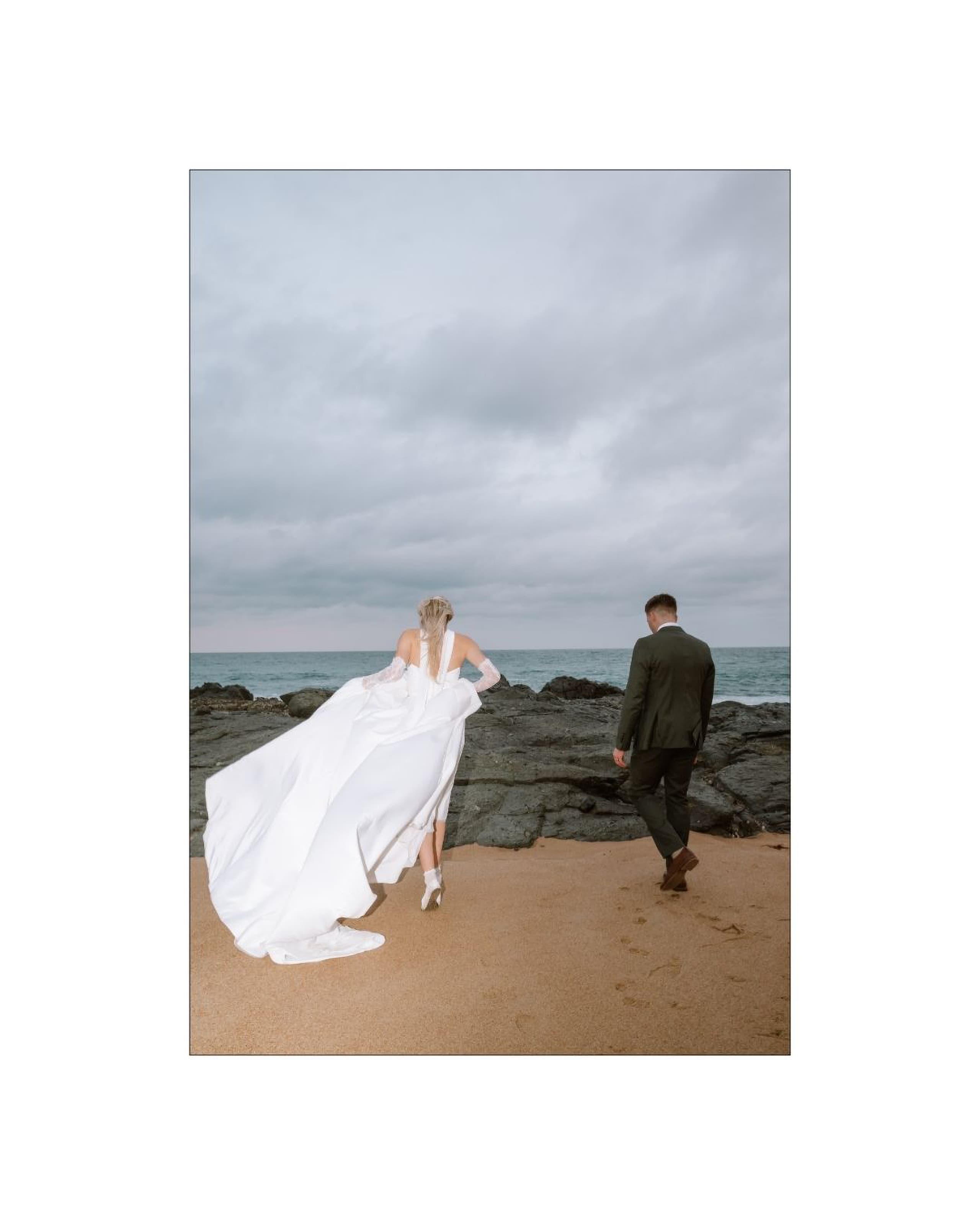 A bride in a flowing white gown walks with the groom across a sandy beach