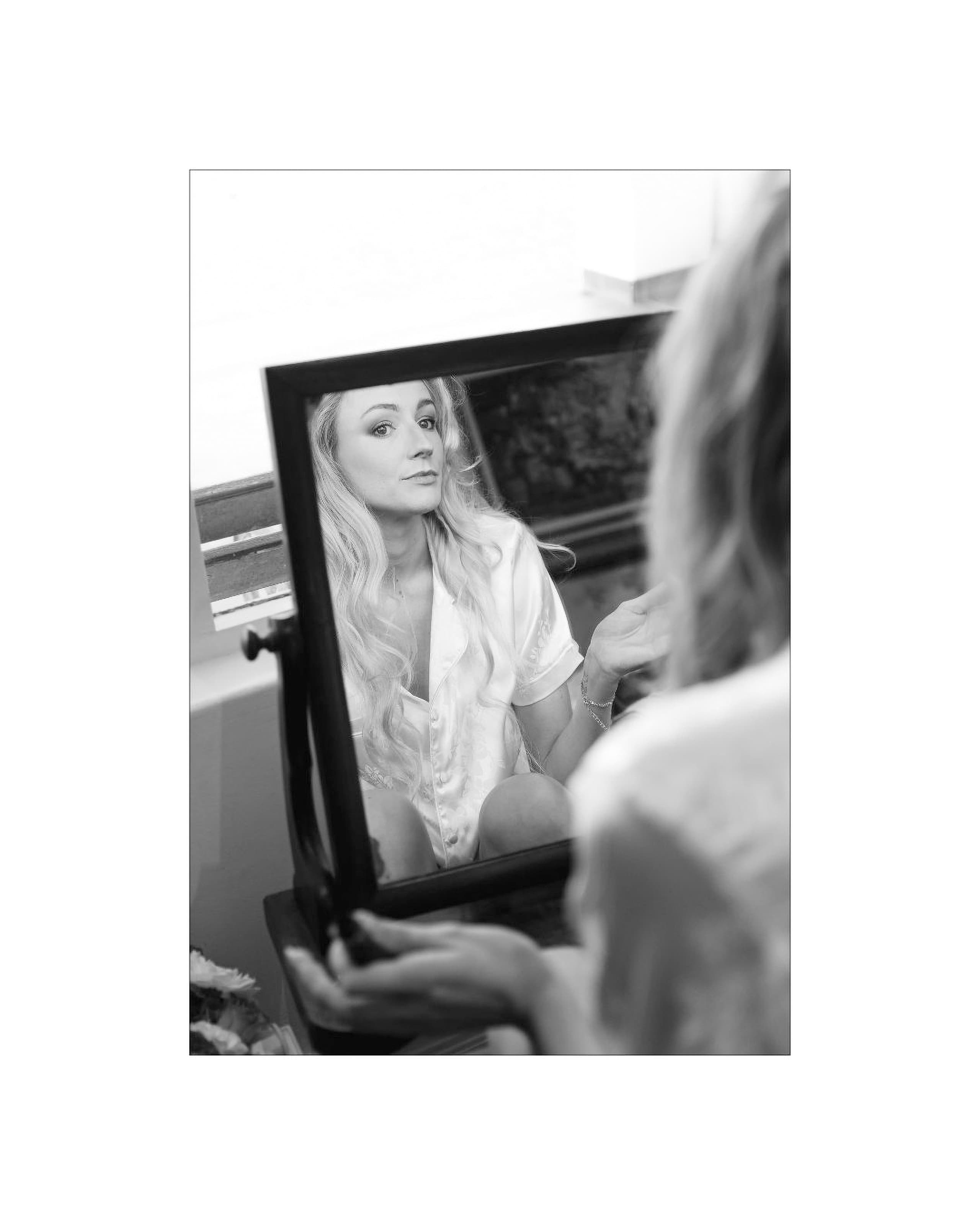A bride with long blonde hair looks into a vanity mirror while getting ready