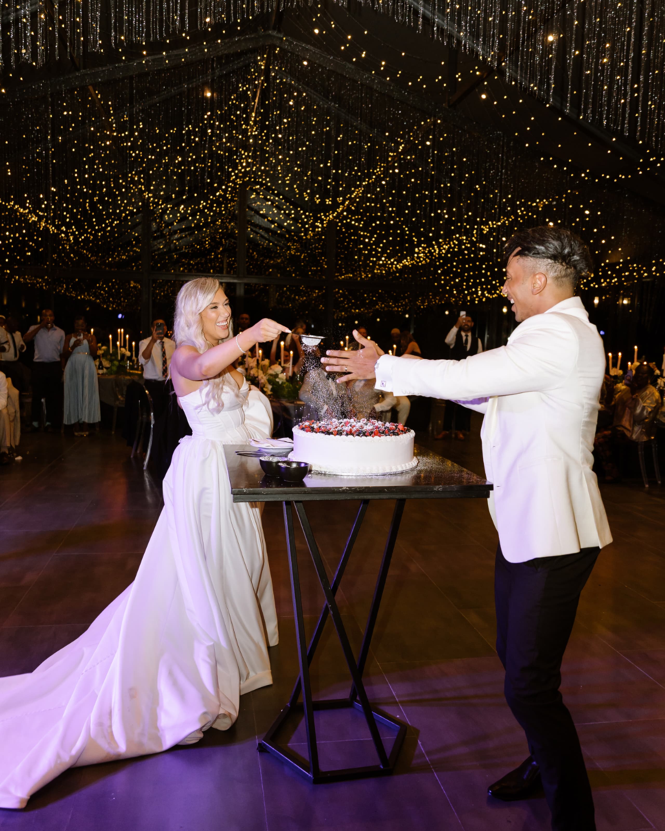 A bride sprinkles powdered sugar over a berry wedding cake while her groom watches happily