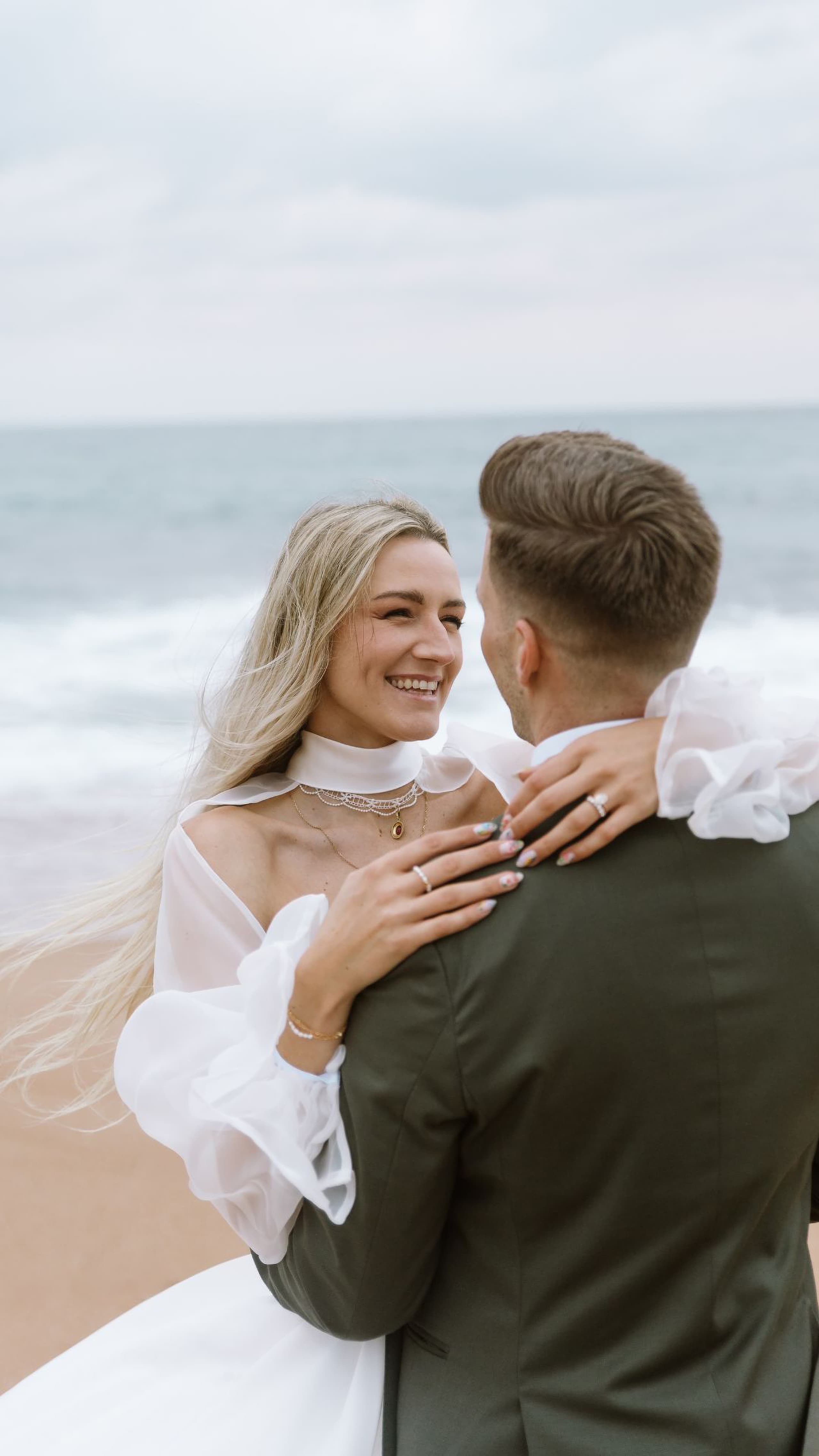 A bride in a white gown and groom in a dark suit embrace on a sandy beach