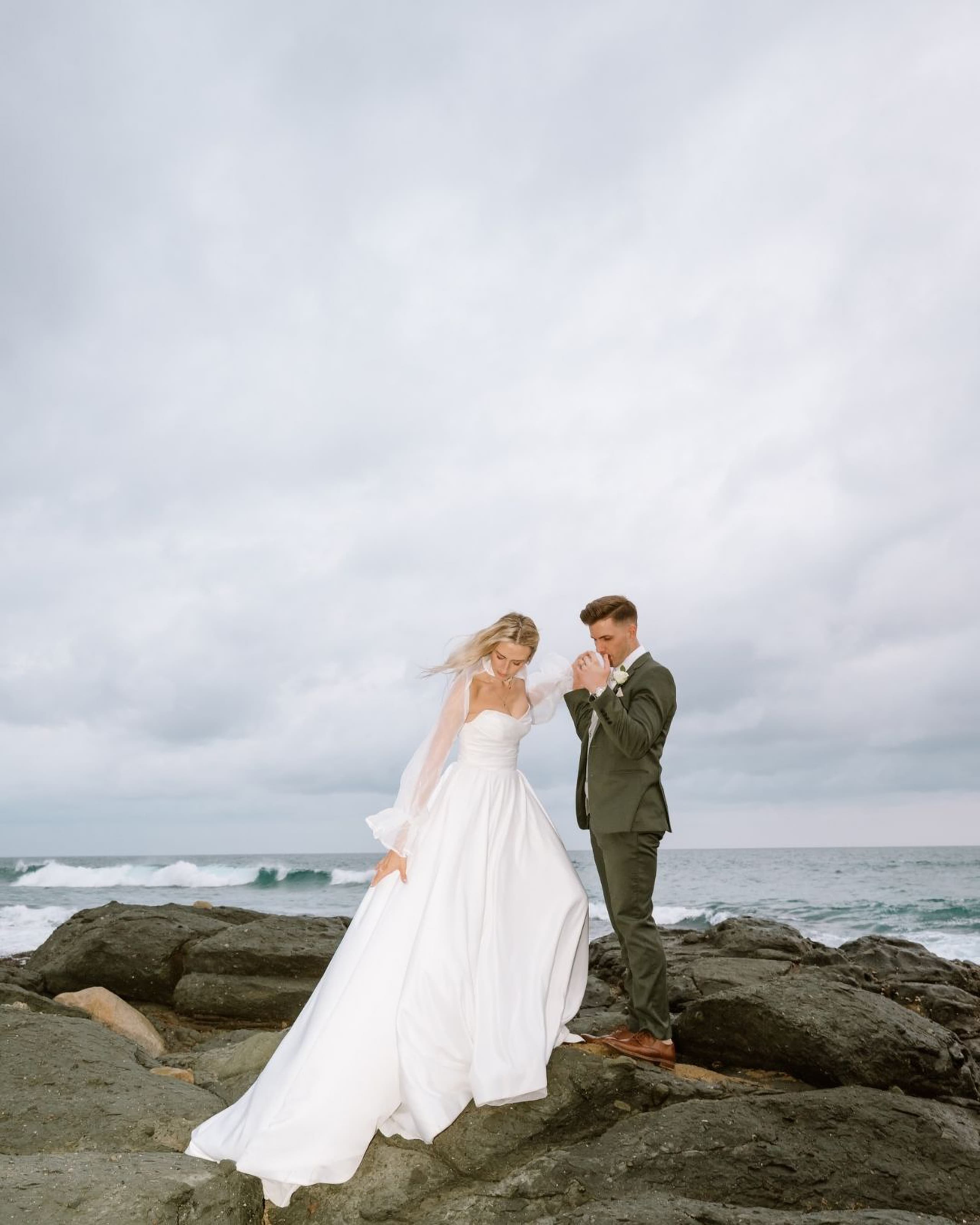 A bride in a white gown and groom in a green suit on coastal rocks