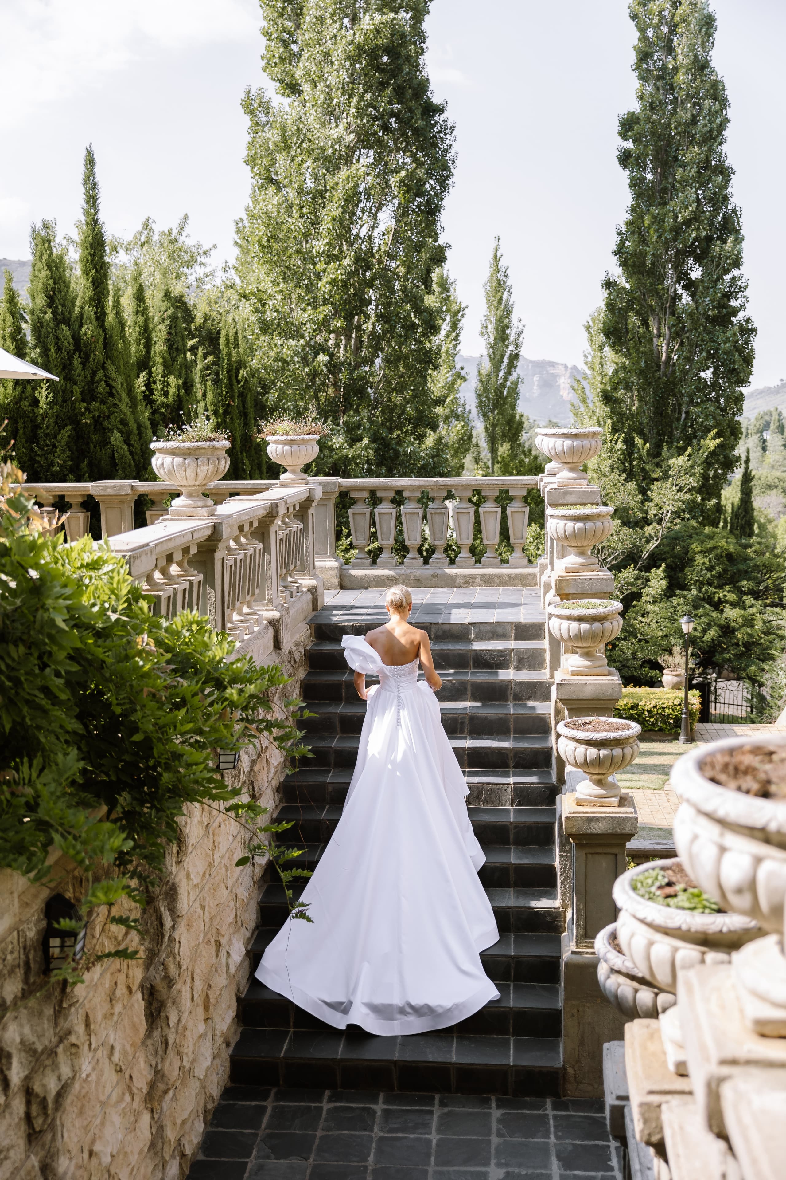 A bride in a white gown walks up stone stairs in a South African garden
