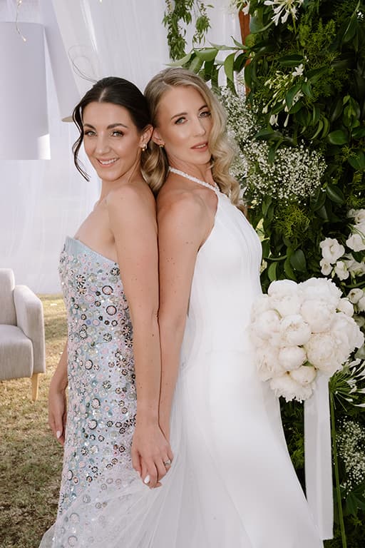 Two women stand back to back in elegant wedding attire amidst lush floral wedding decor