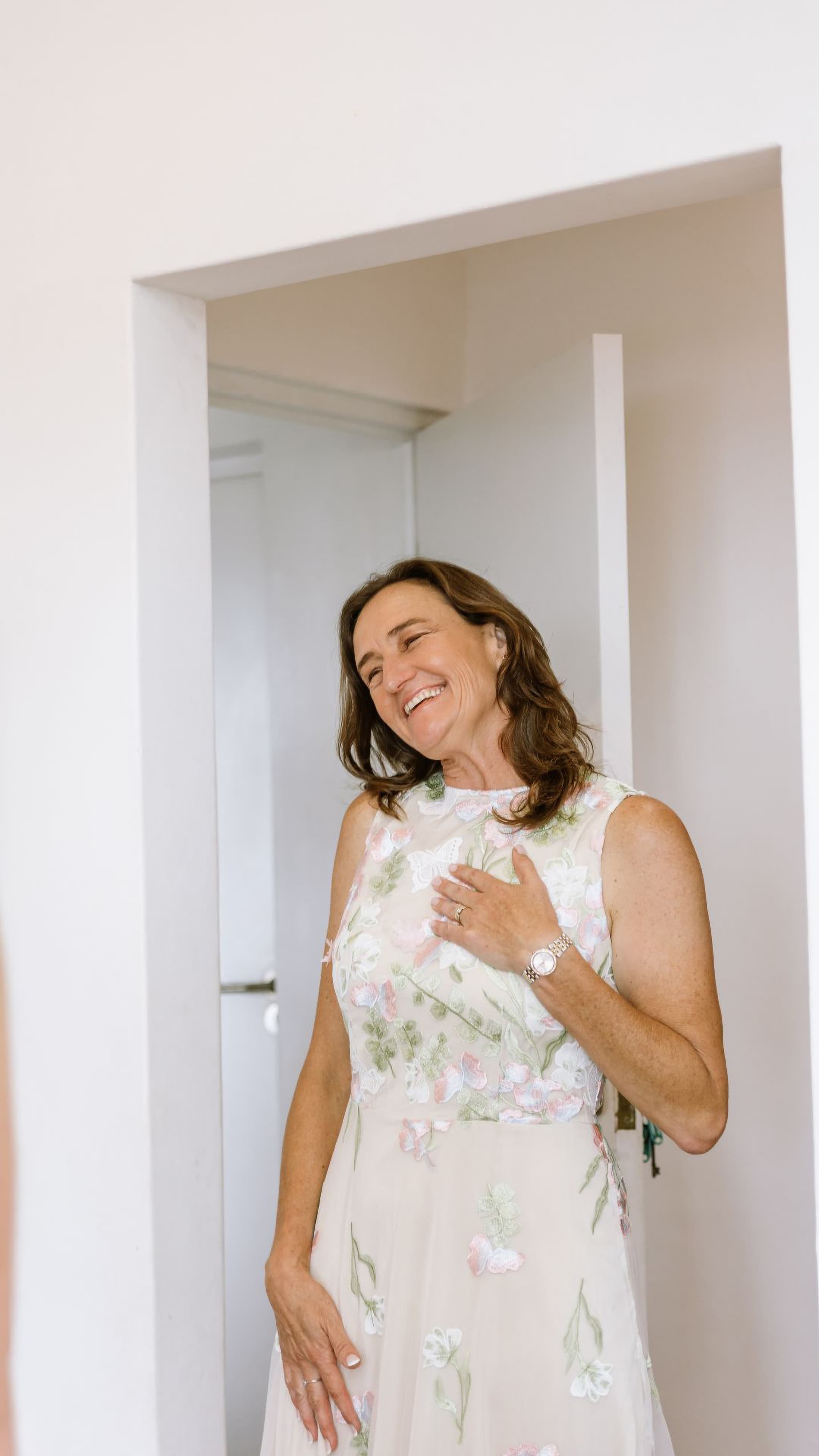 An emotional mother of the bride smiling joyfully with her hand over her heart indoors