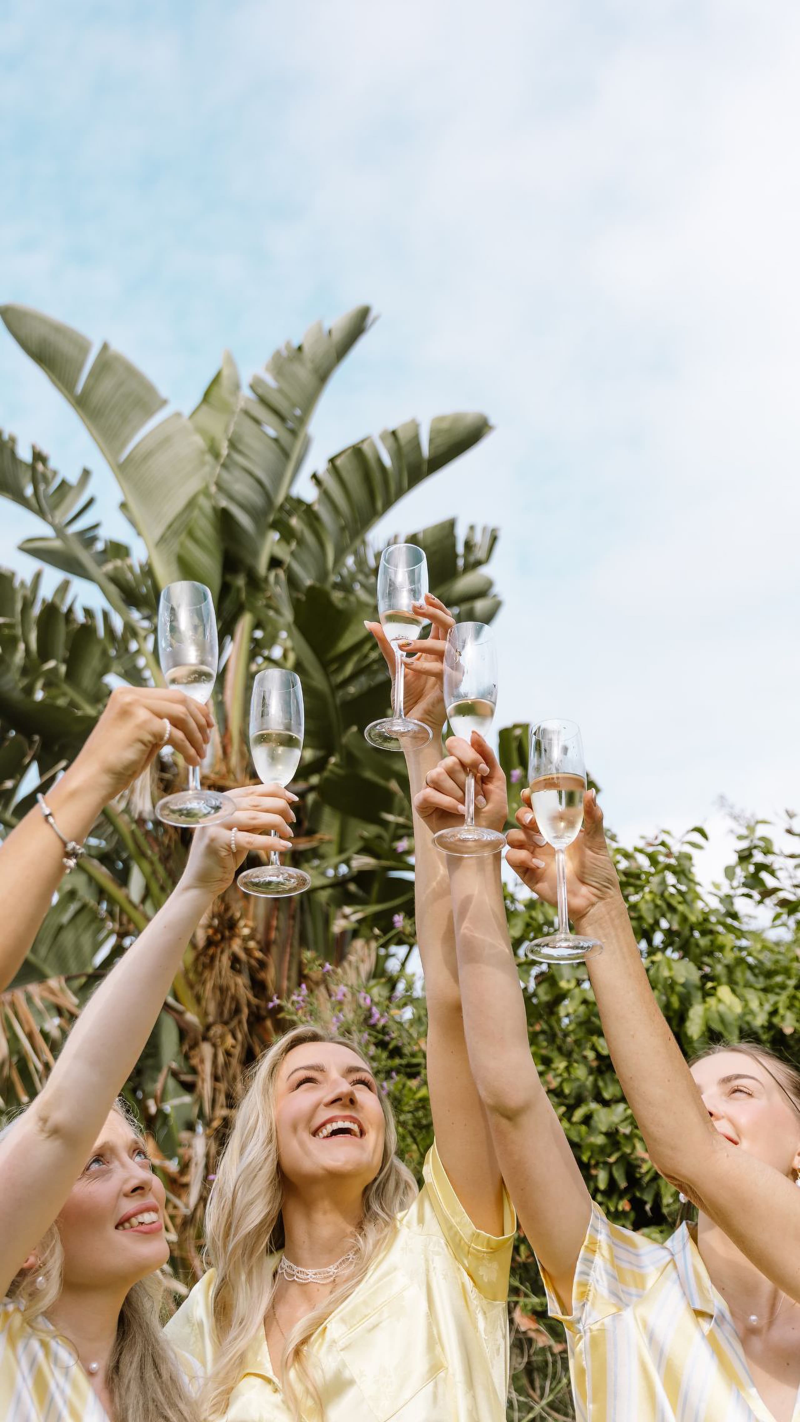 Four bridesmaids in yellow pajamas raising champagne flutes outdoors under blue skies