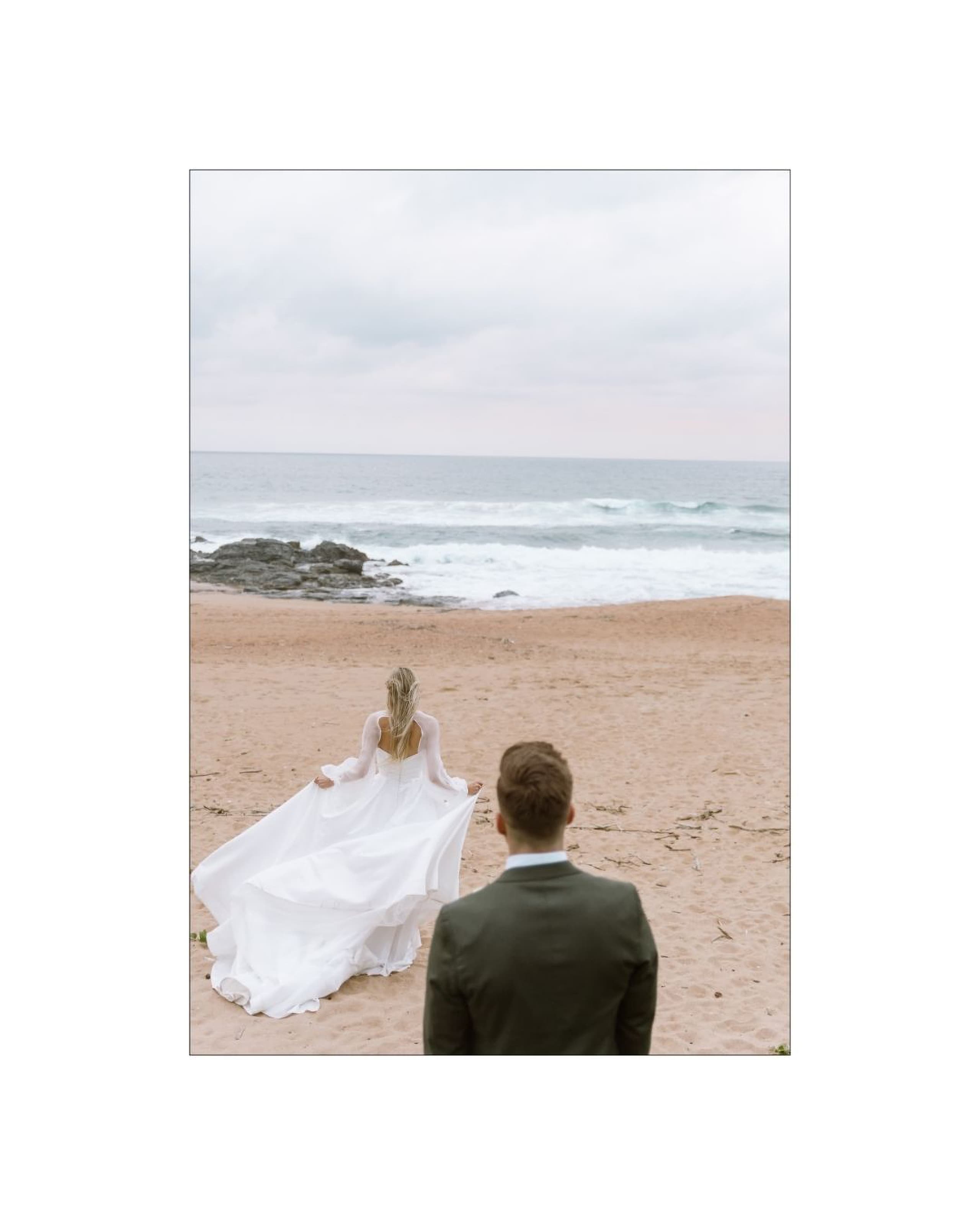 A groom in a dark suit watches his bride in a flowing white dress walk toward the ocean on a sandy beach