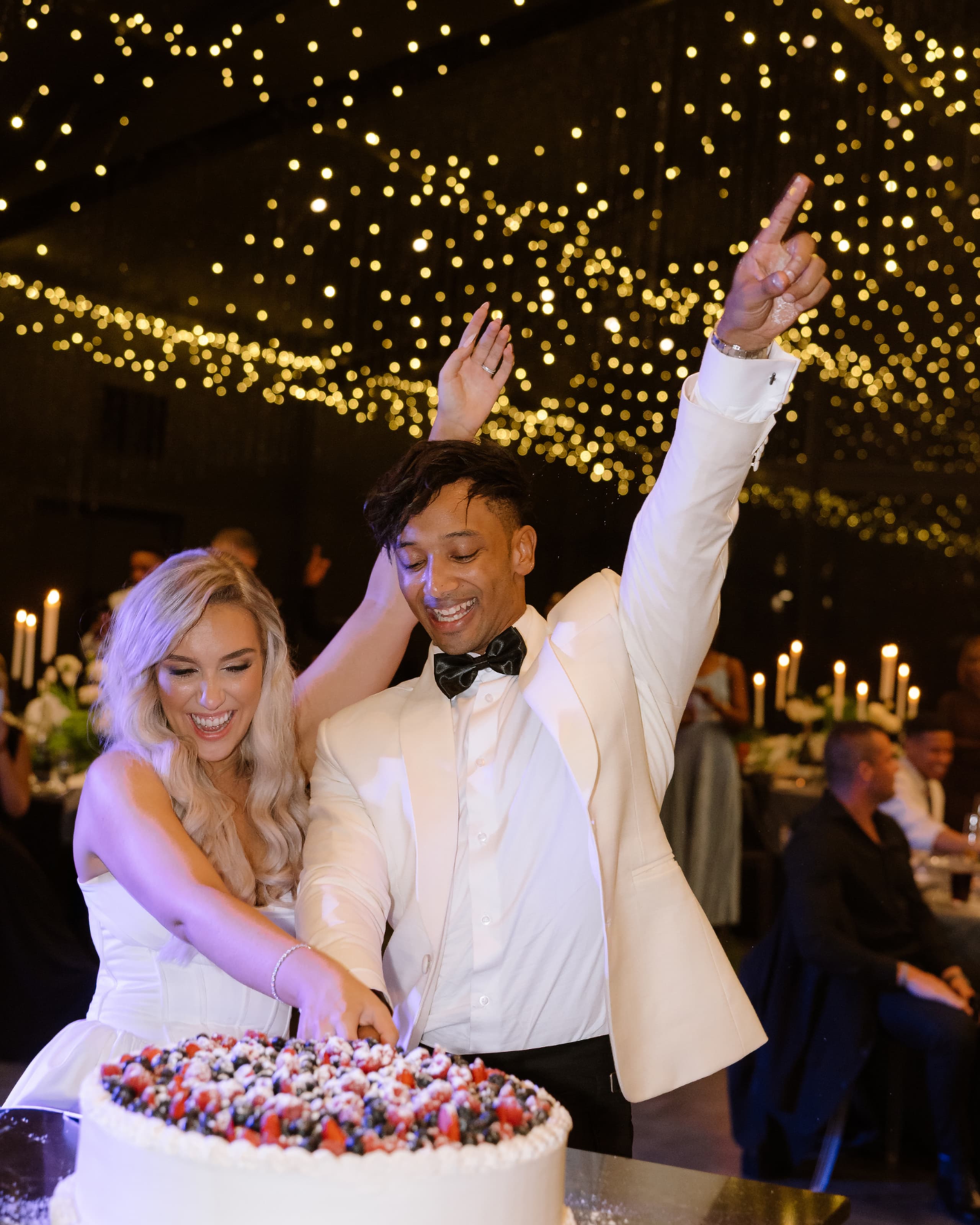A joyful bride and groom cut their berry-topped wedding cake under sparkling fairy lights