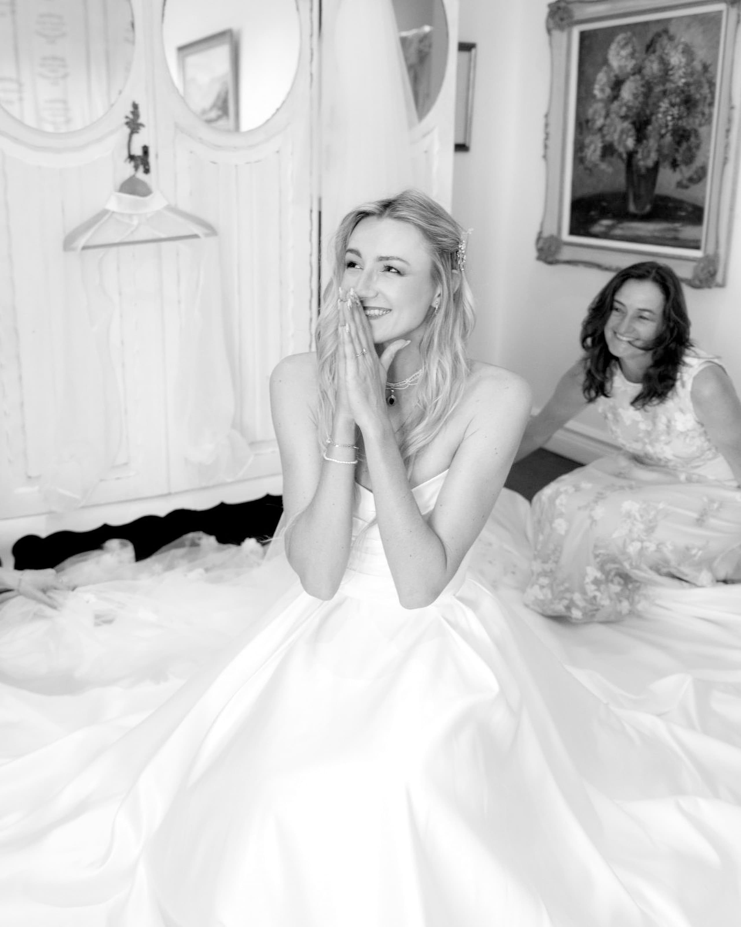 A joyful bride smiles with her hands clasped while sitting in her wedding dress
