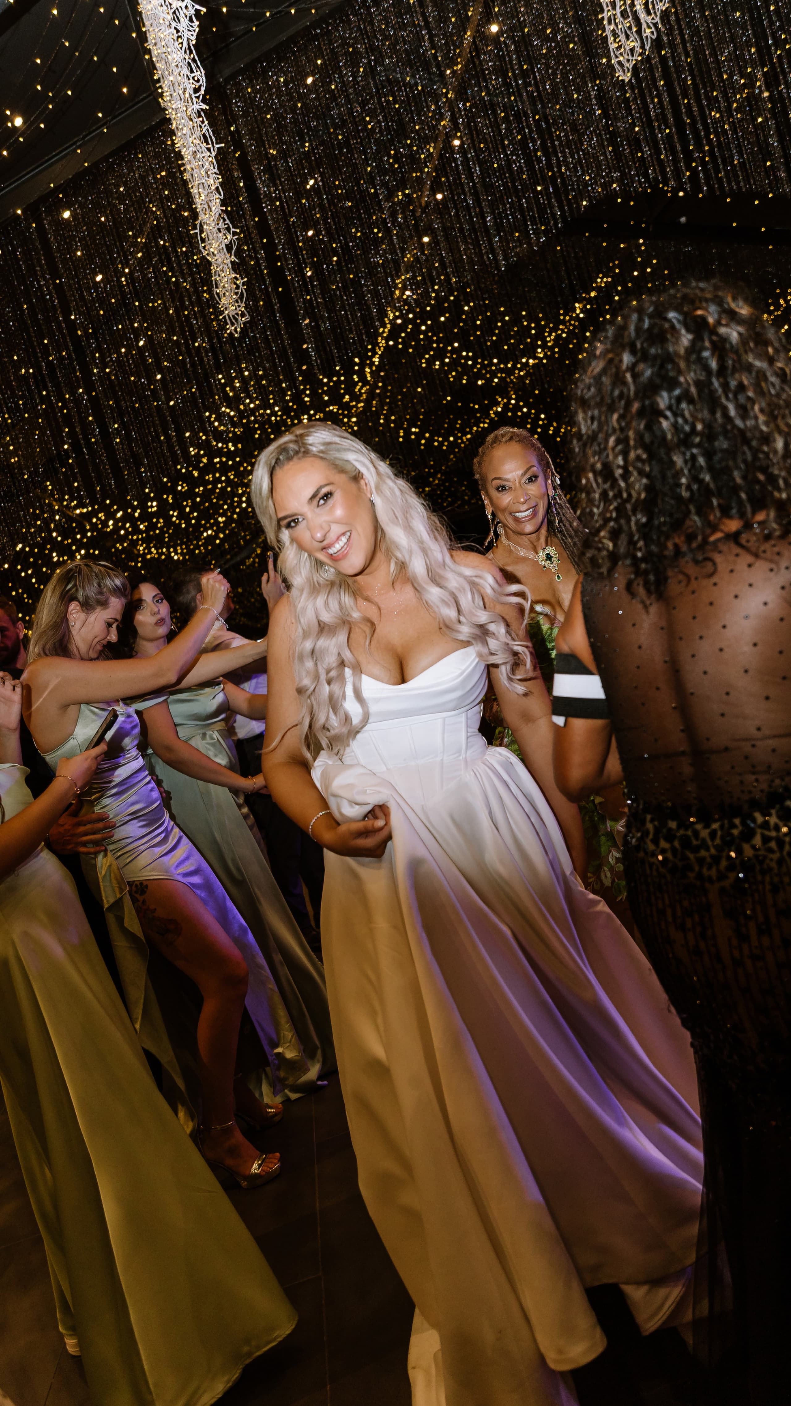 A joyful bride in a white corset gown dancing with guests under twinkling fairy lights