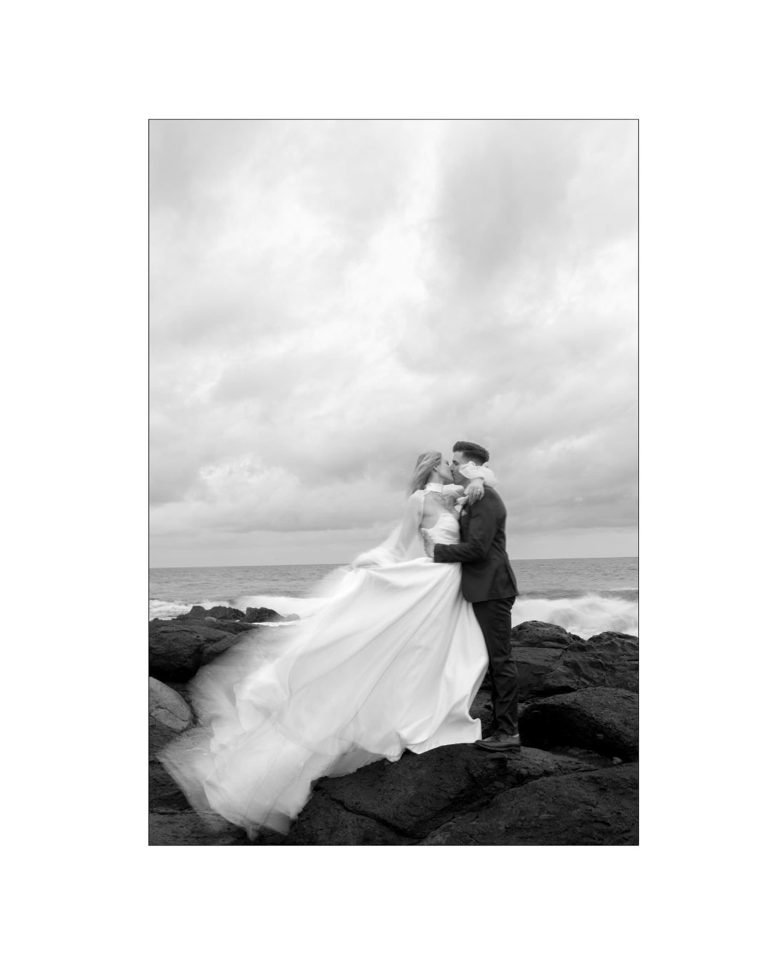 Newlyweds kiss passionately on dark coastal rocks beneath a dramatic overcast sky