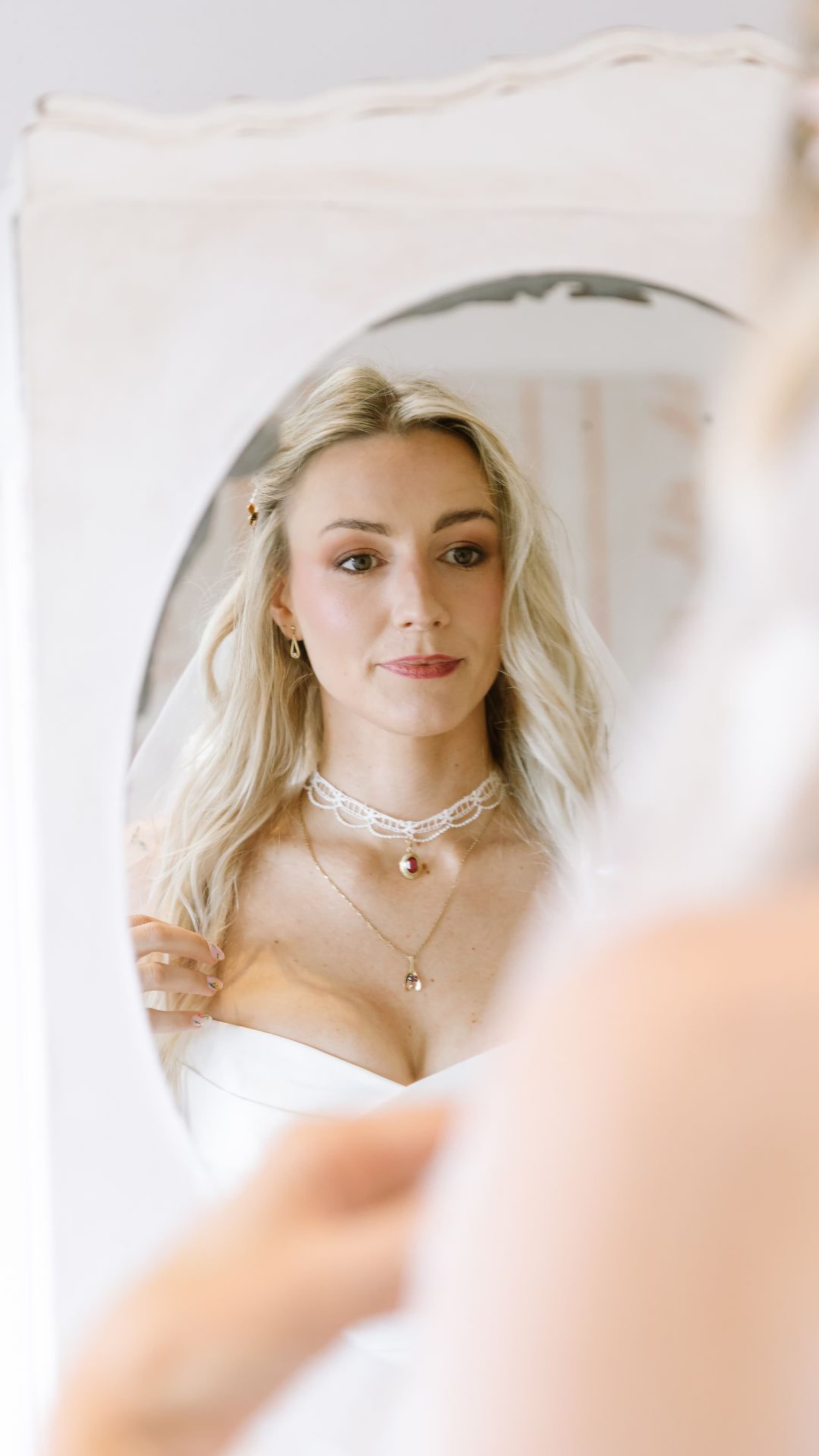A radiant blonde bride adjusts her white wedding dress while looking into an oval mirror