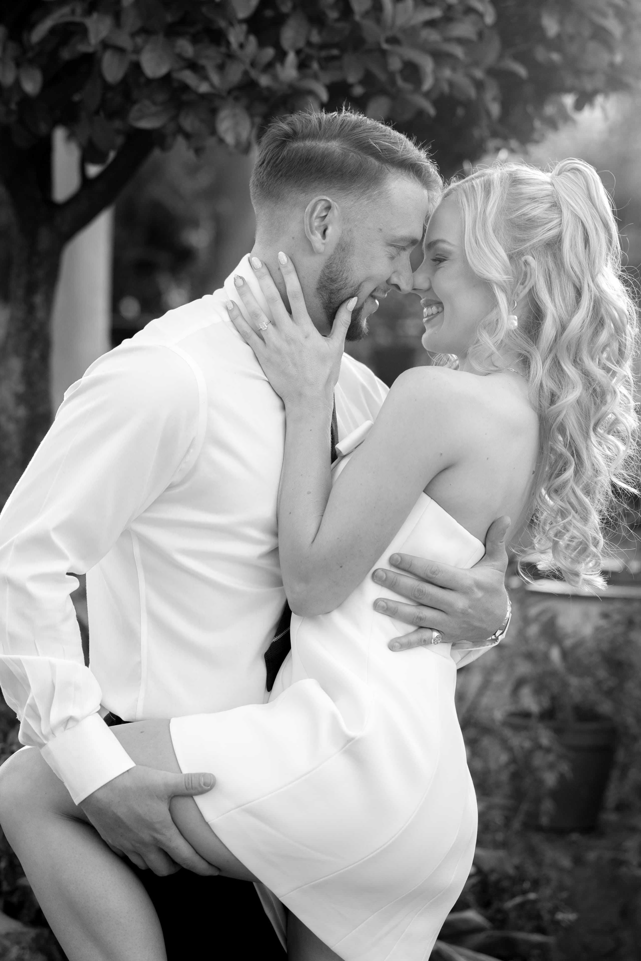 A romantic couple in formal attire embraces intimately while smiling at each other outdoors