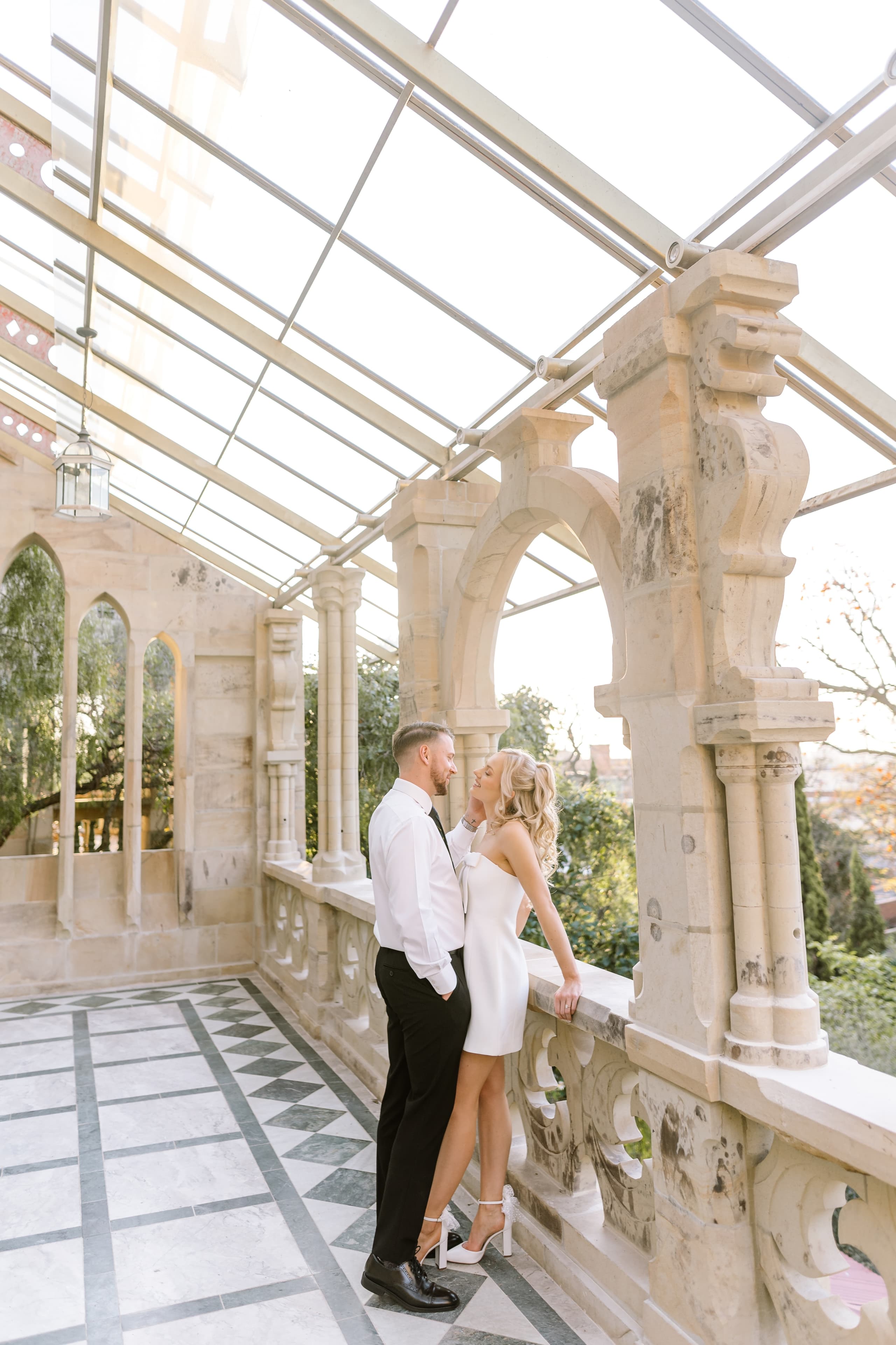 A newlywed couple shares a romantic moment on a sunlit stone terrace with glass ceilings