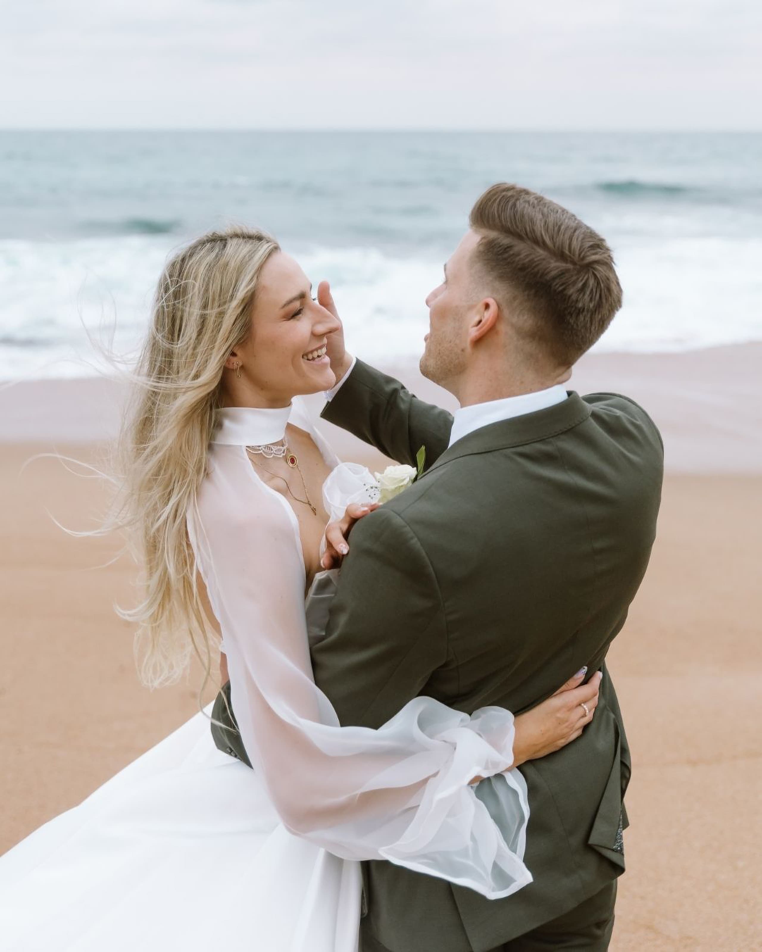 A smiling bride and groom embrace joyfully on a sandy beach near the ocean