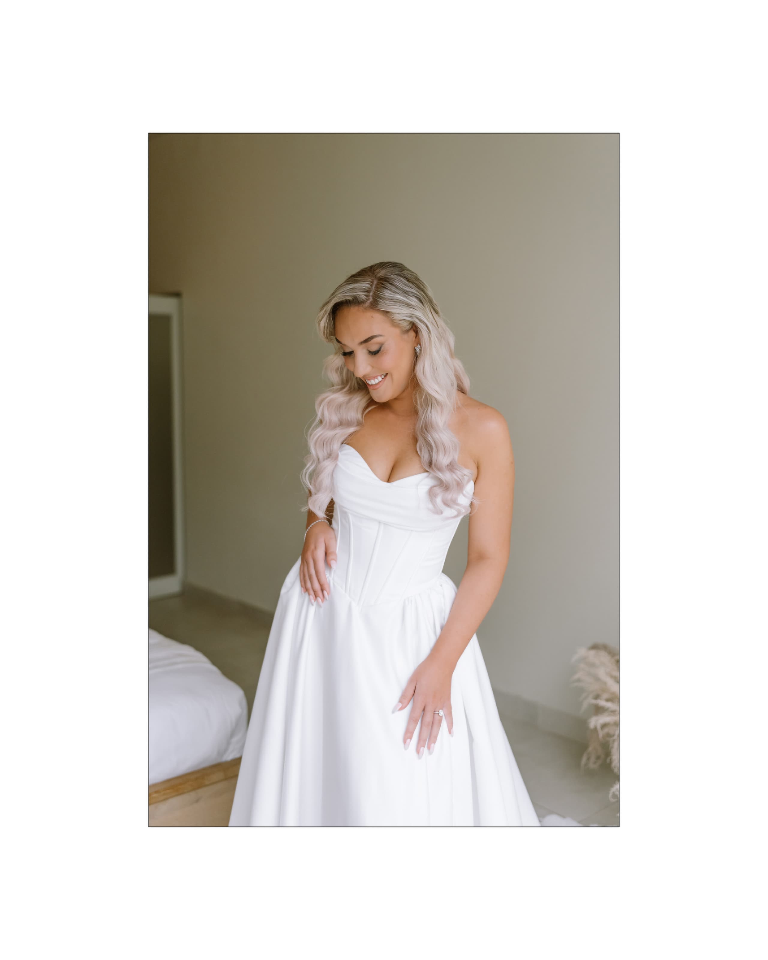 A smiling bride wearing a white strapless corset wedding dress stands in a bright room