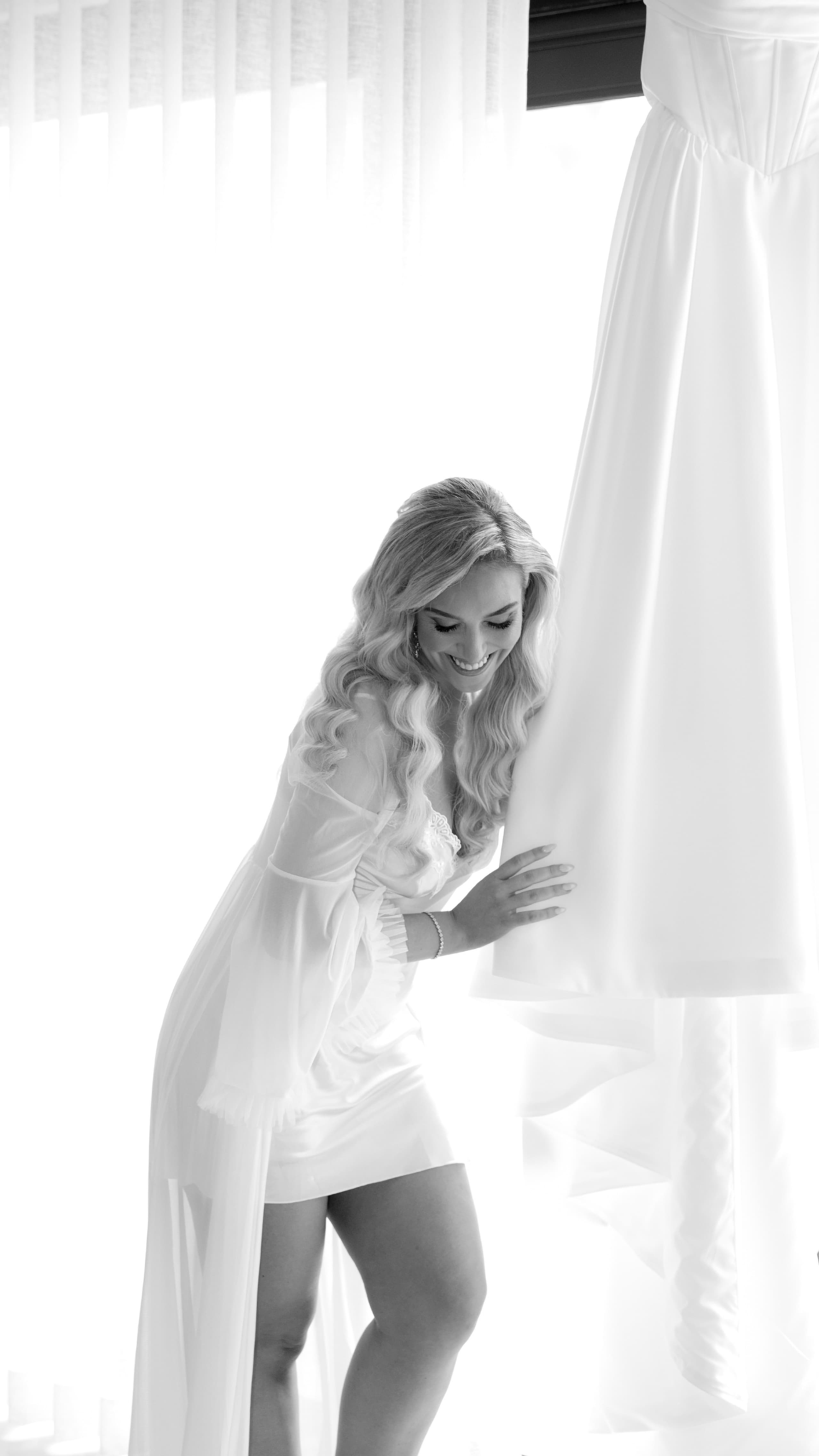 A smiling bride in a white silk robe touches her hanging wedding dress near window light