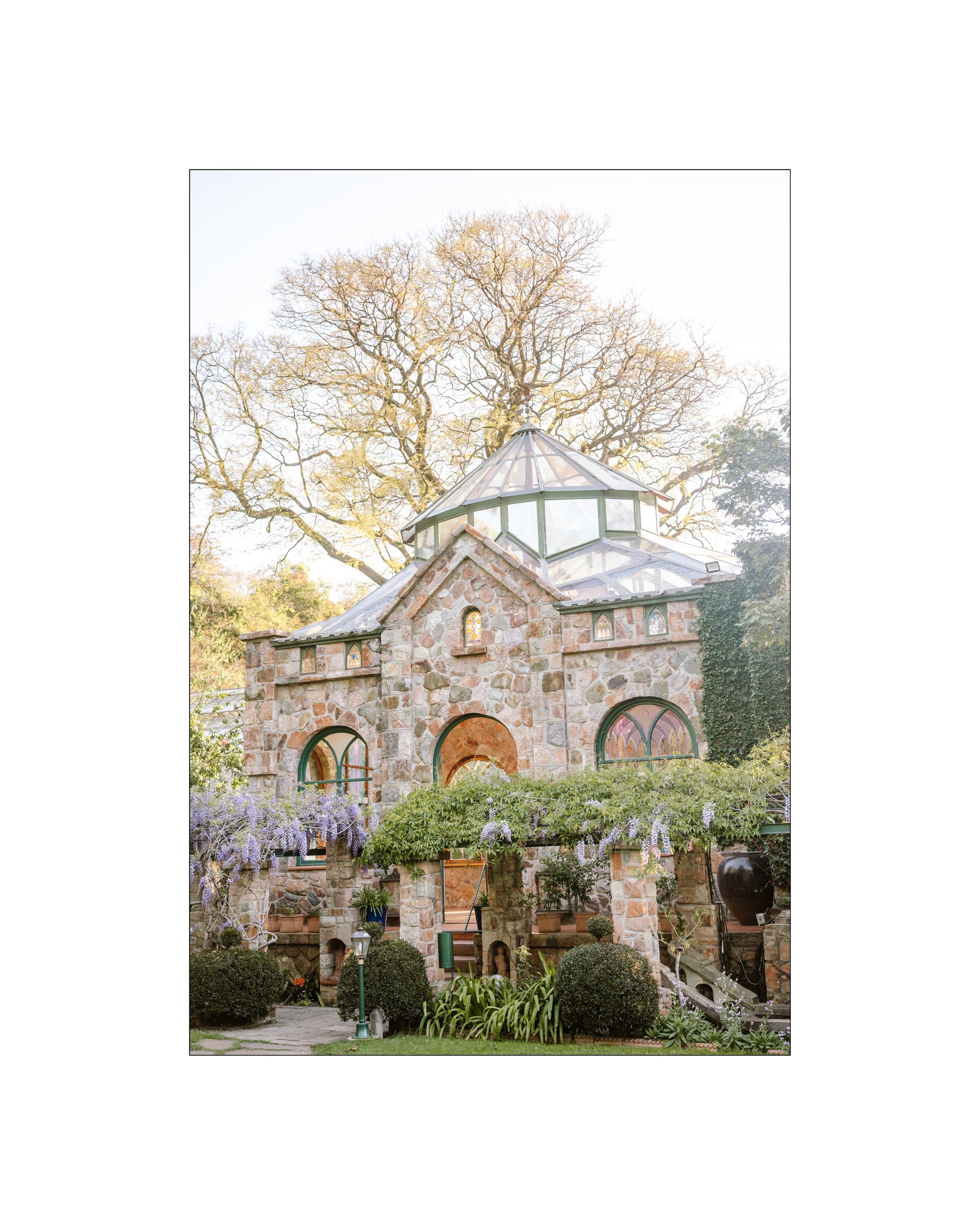 A stone conservatory with a glass dome roof stands surrounded by blooming purple wisteria vines