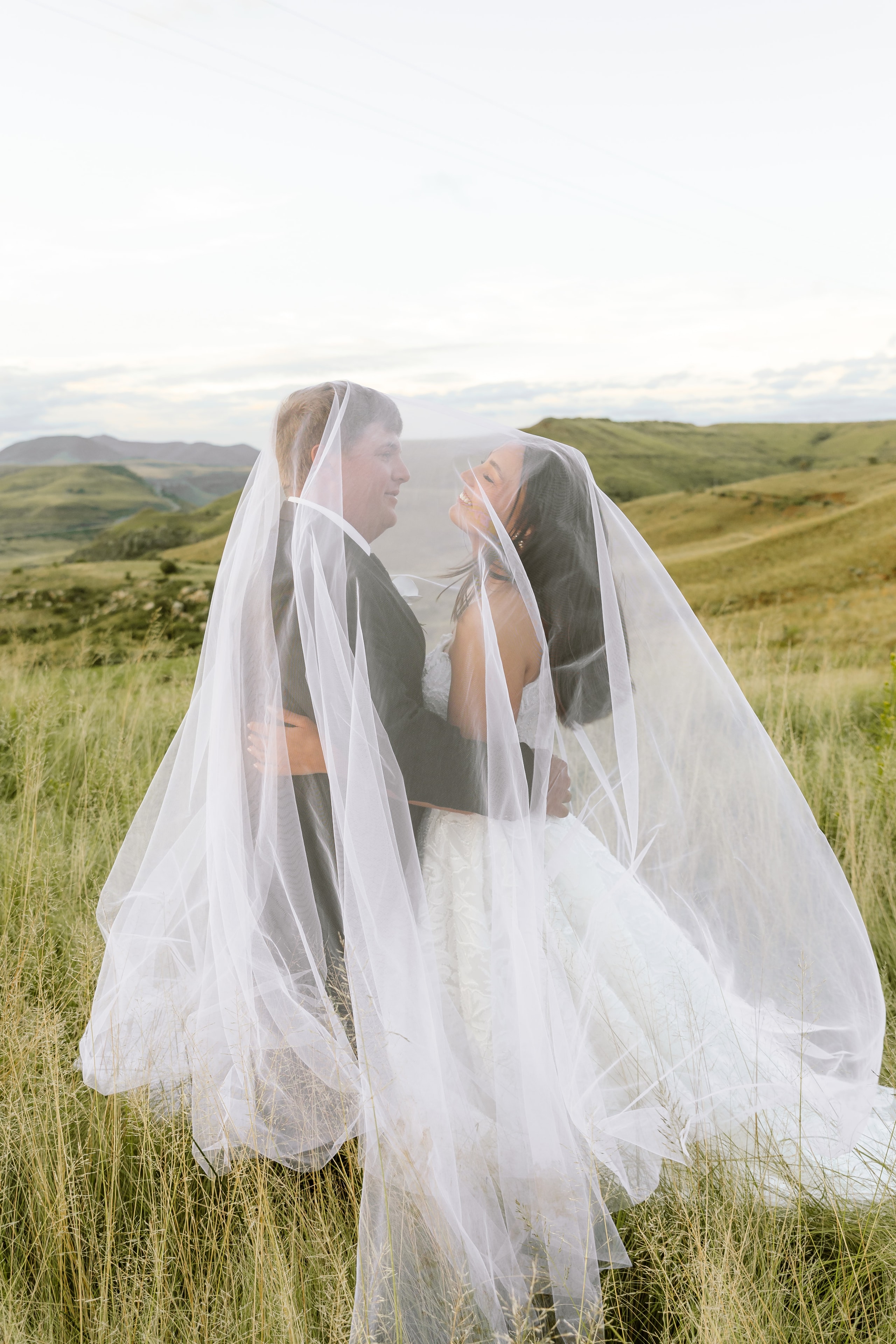 A bride and groom share a joyful moment underneath a long white veil outdoors