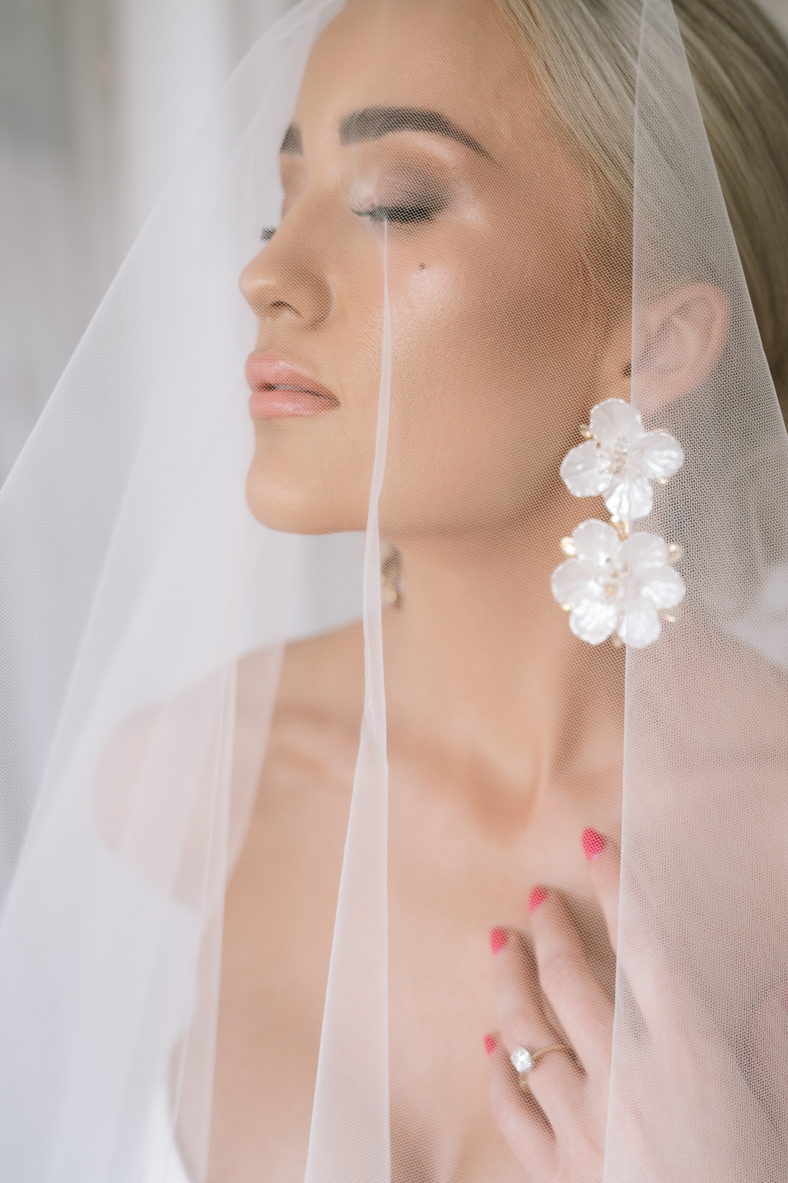 A bride wearing a delicate veil and floral earrings poses with her hand on her chest