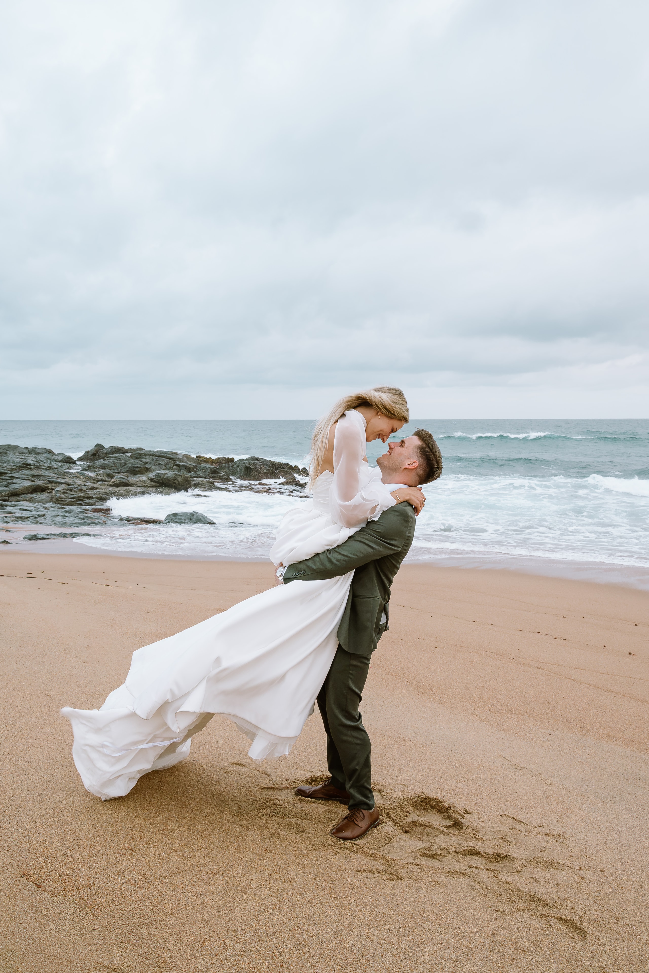 A groom in an olive suit lifts his bride in a flowing white gown on a beach