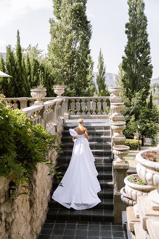 A bride in a satin gown walks up stone garden stairs lined with urns and cypress trees
