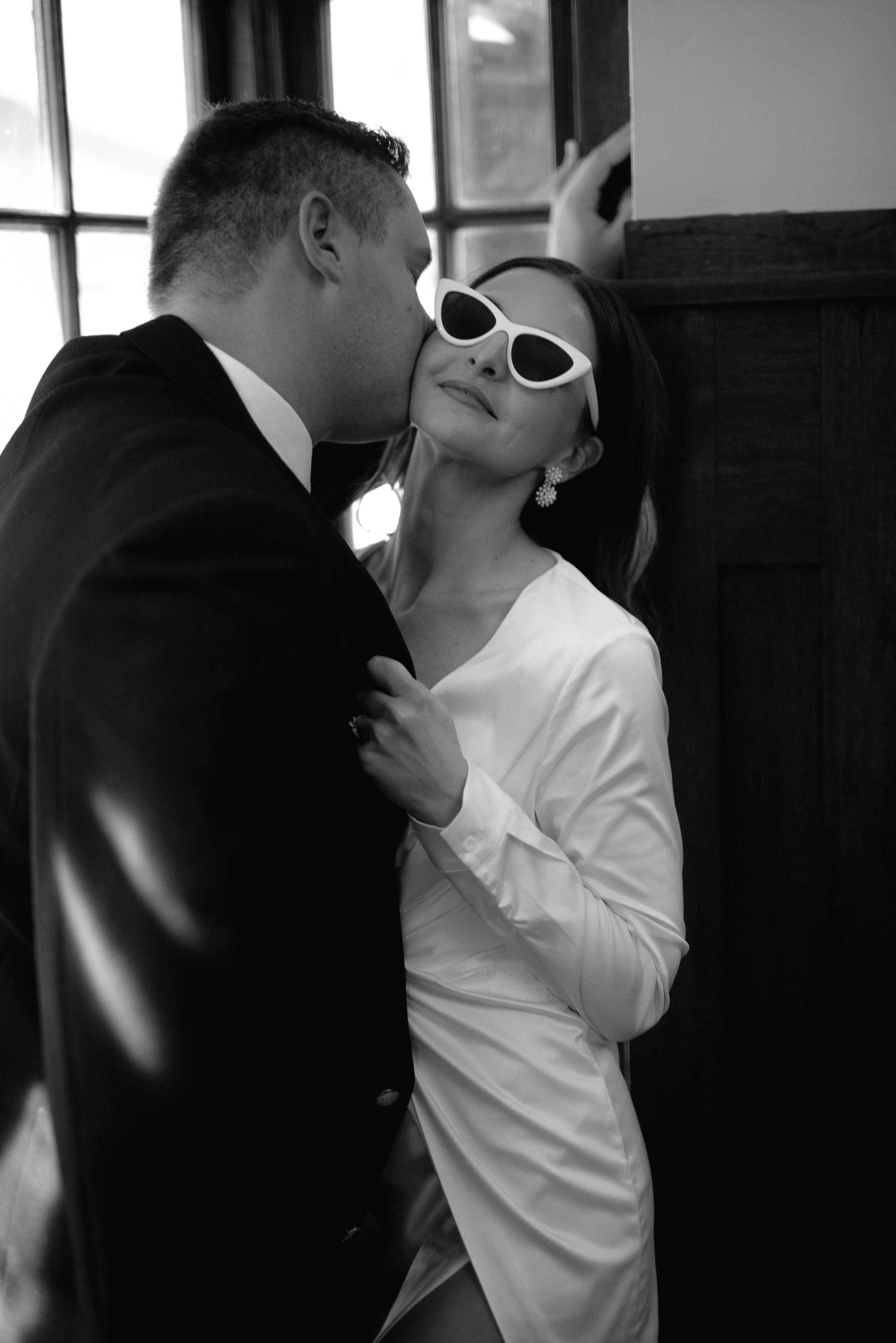 A groom kisses his bride on the cheek while she wears stylish white cat-eye sunglasses