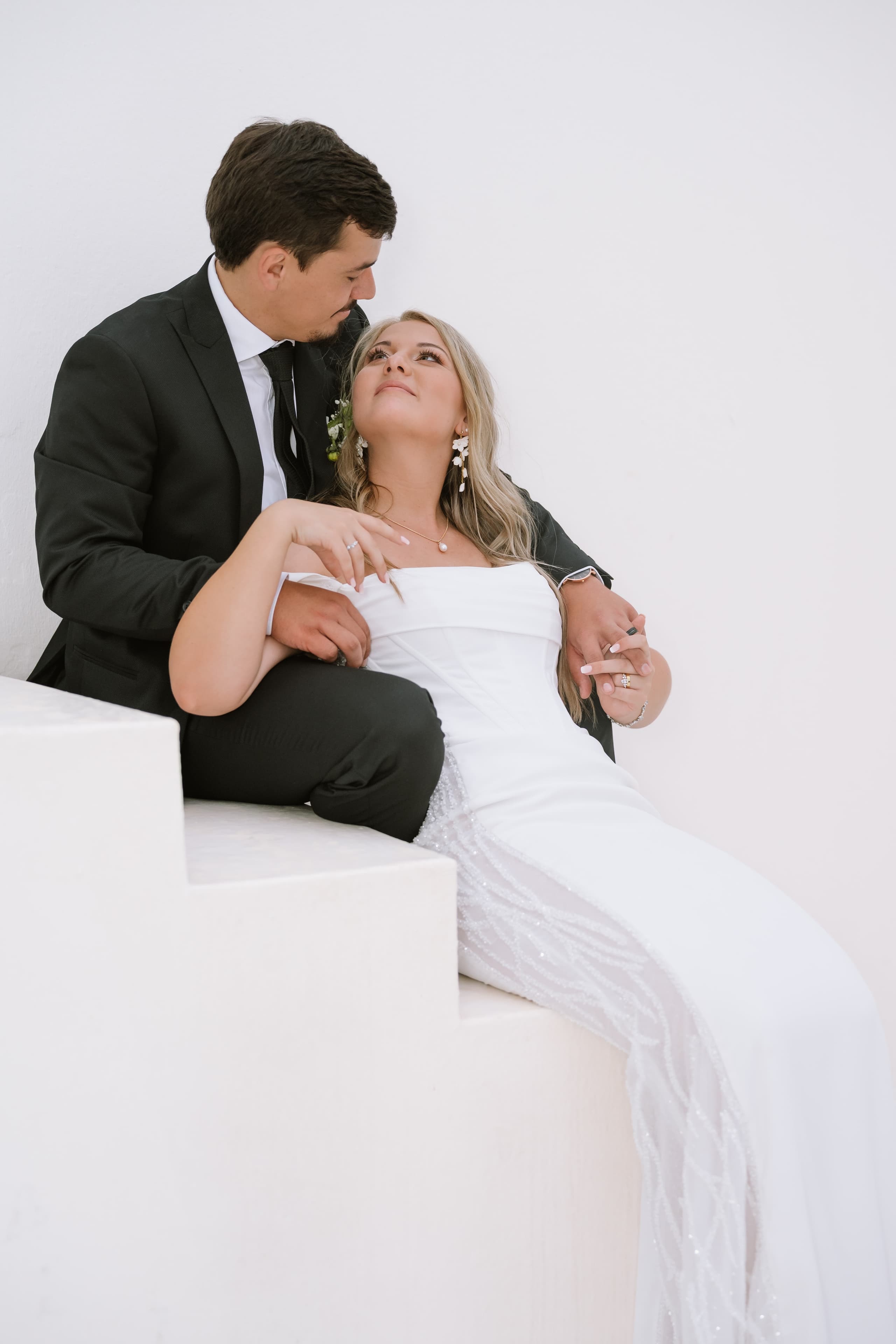 A bride and groom sit together on white stairs against a minimalist white background