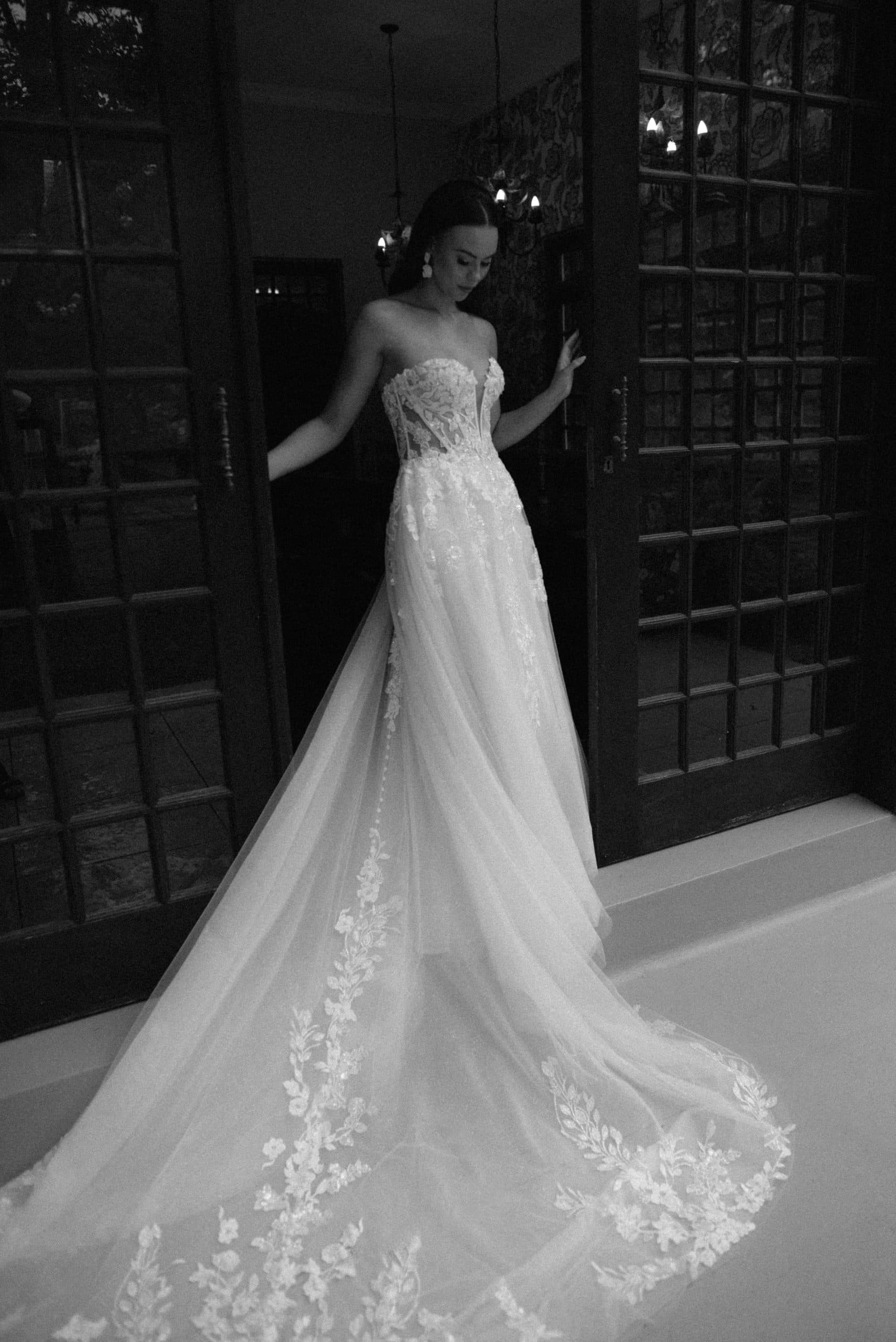 A bride in a strapless lace wedding dress stands gracefully in a dark doorway