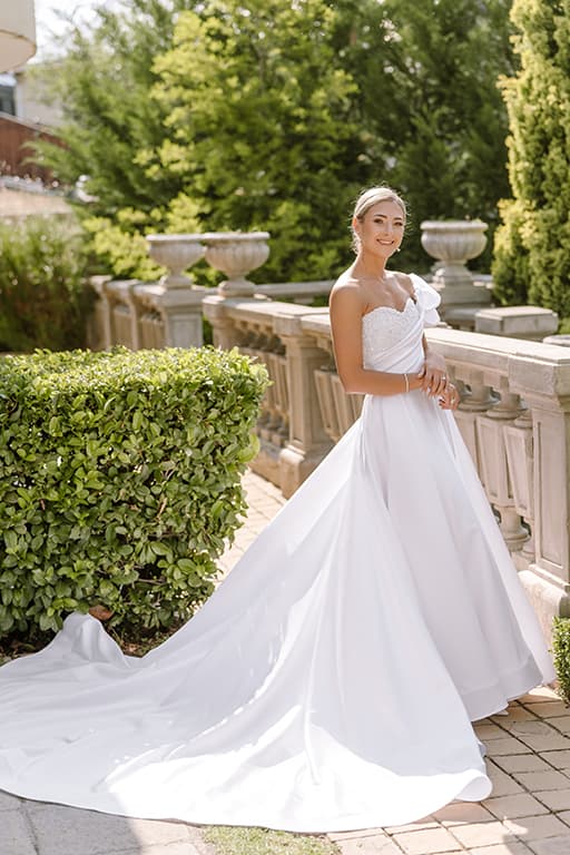 A bride in a strapless satin gown with a long train beside a stone garden balustrade