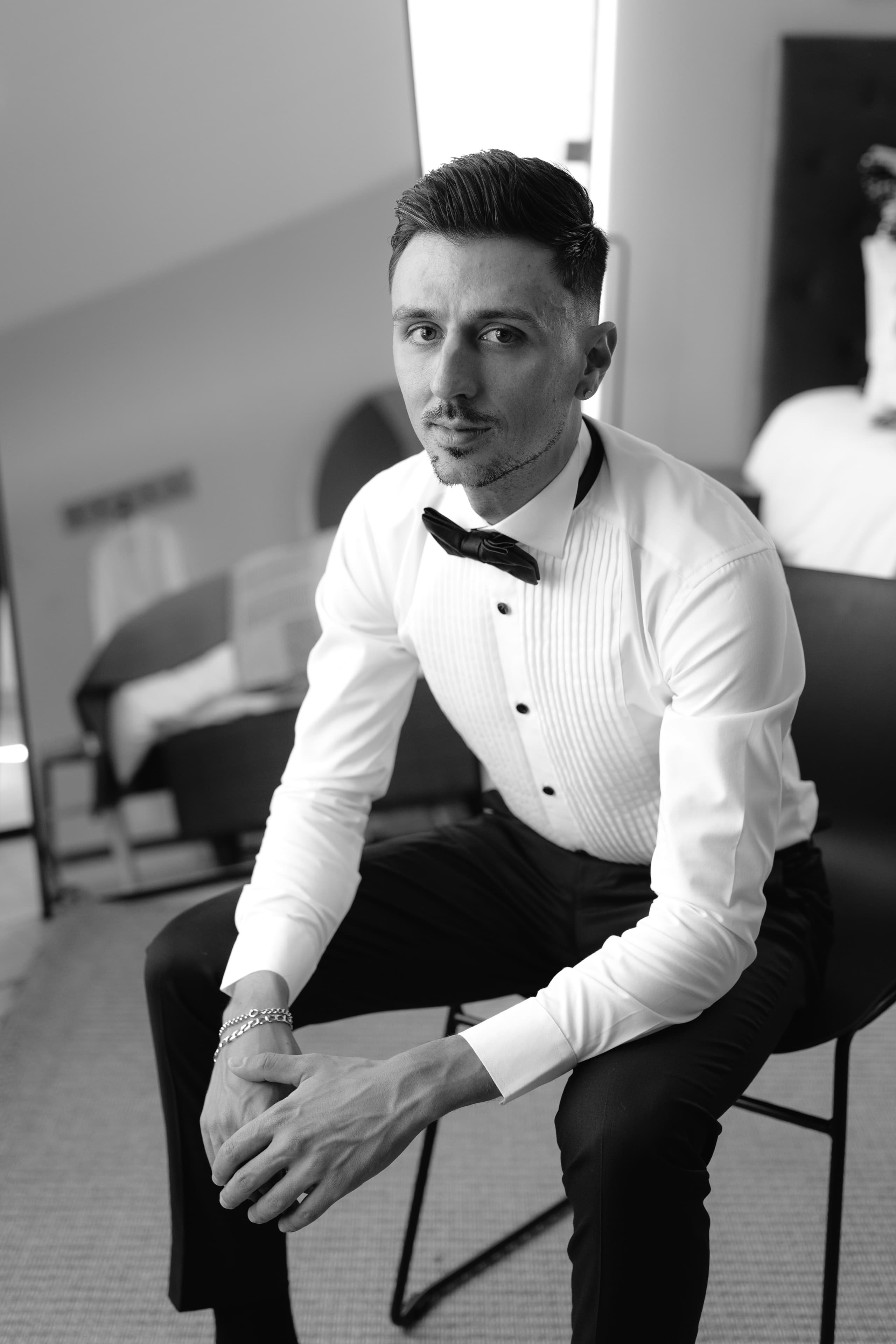 A stylish groom in a crisp white tuxedo shirt and black bowtie sits waiting