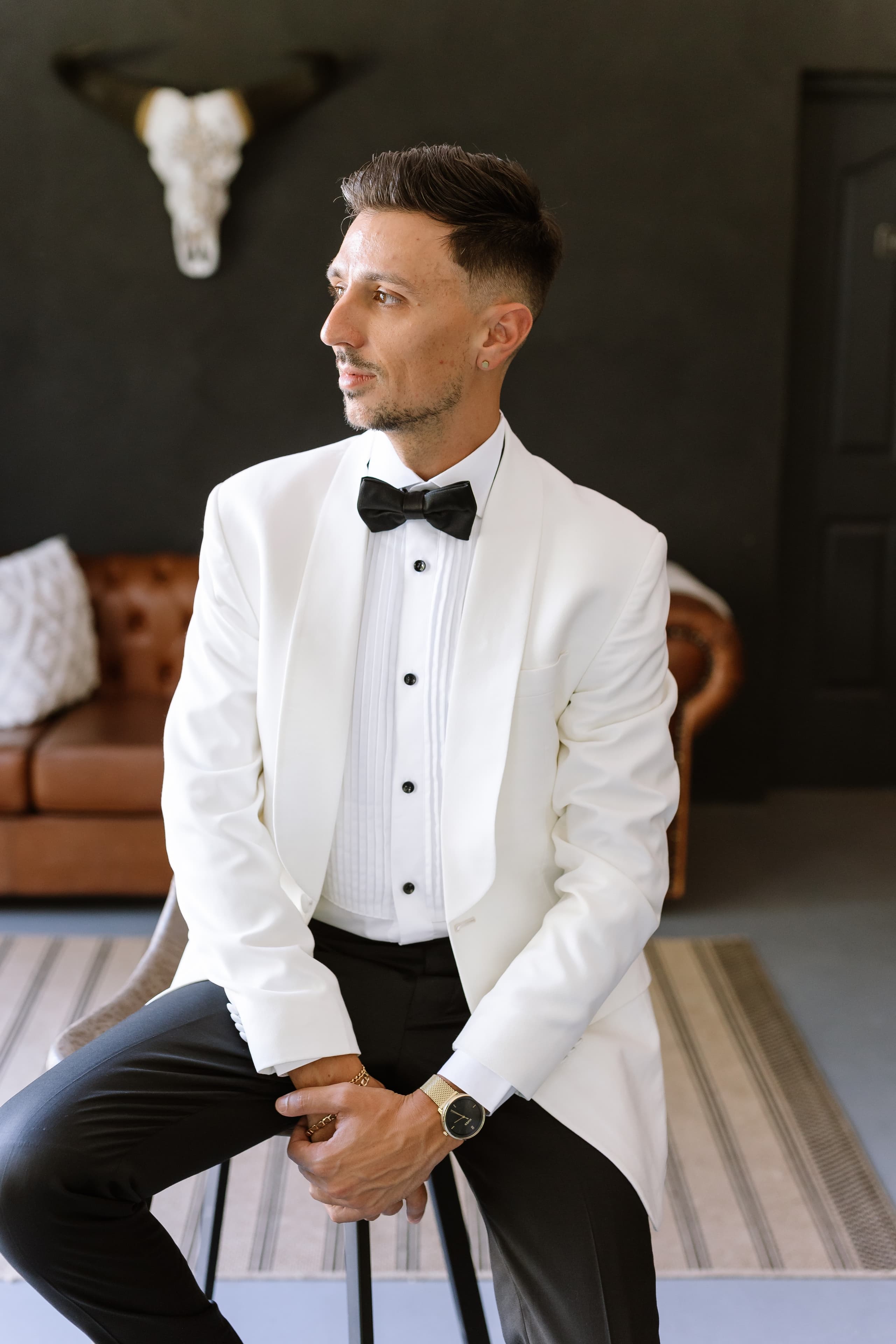 A stylish groom in a white tuxedo jacket and black bow tie sits elegantly indoors