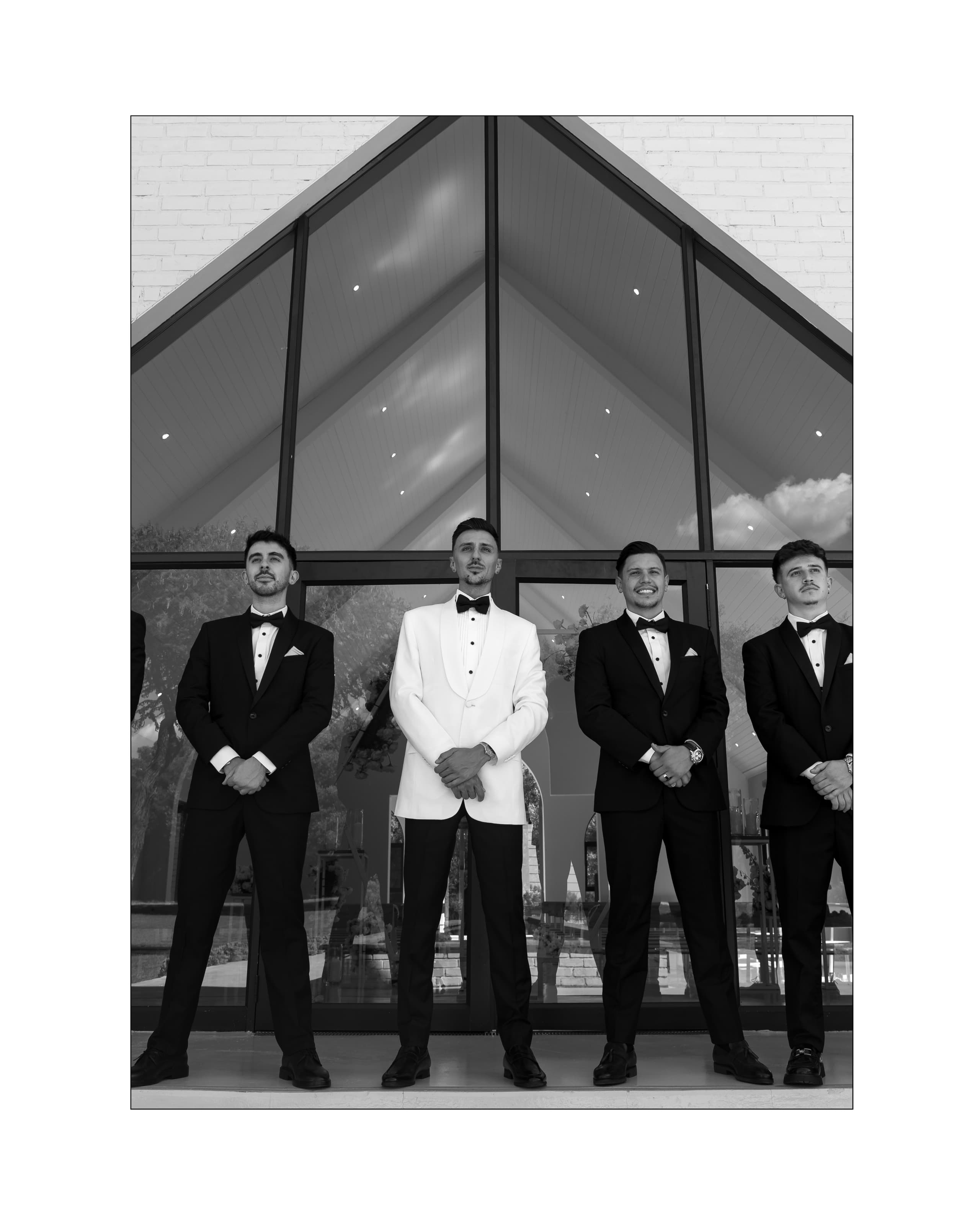 Four men in formal black and white tuxedos stand confidently before a modern glass building
