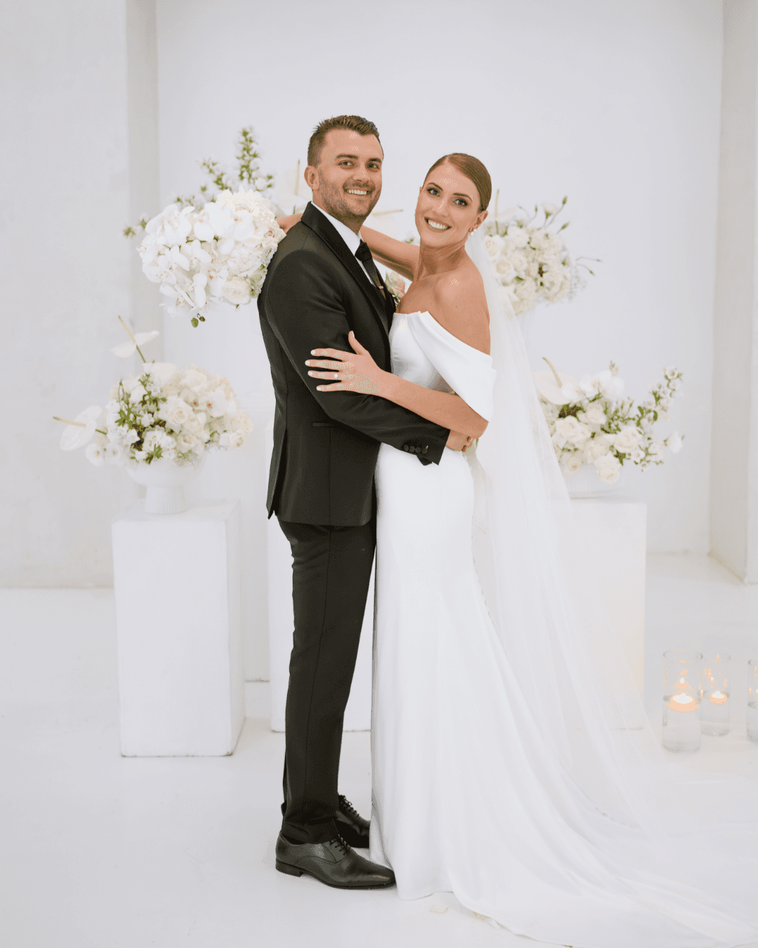 A smiling bride and groom embrace in front of elegant white floral arrangements at their wedding