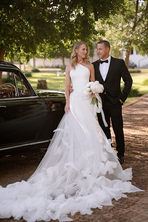 A bride and groom beside a vintage black car under the trees