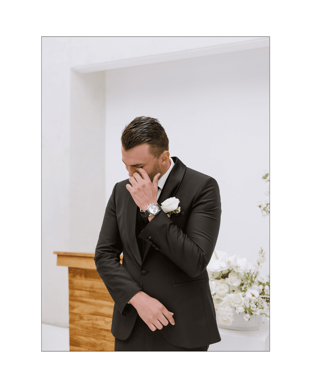 A groom in a black tuxedo wipes away a tear during his wedding ceremony