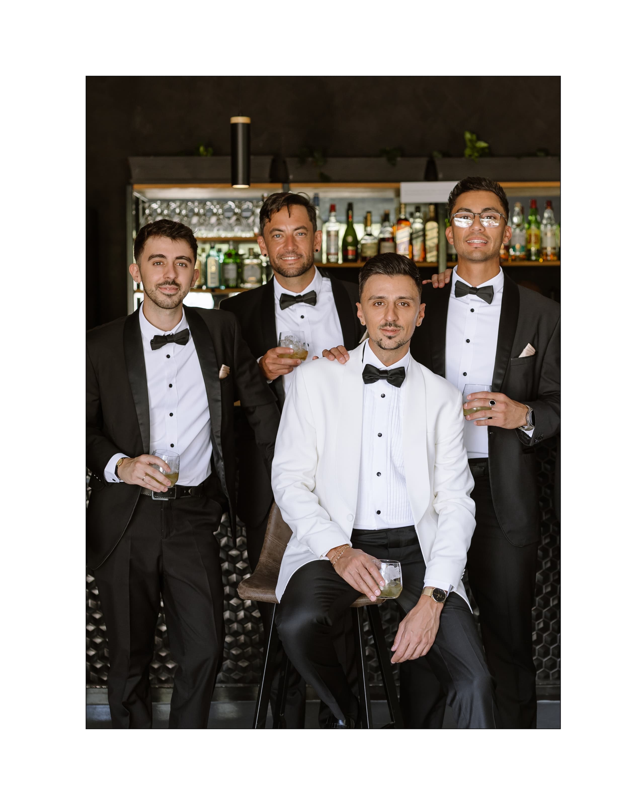 Four groomsmen in elegant black and white tuxedos posing together at a modern bar