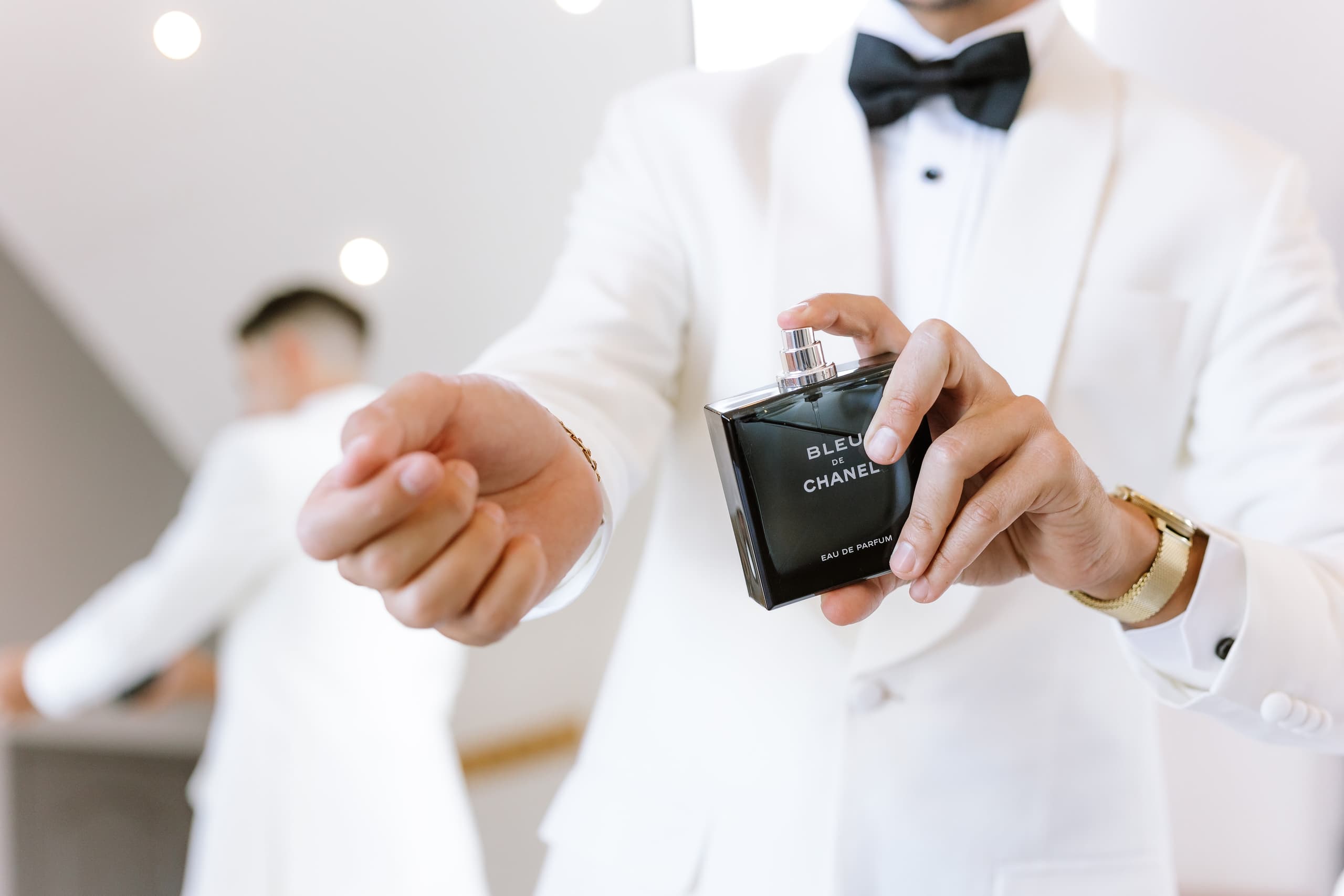 A groom in a white tuxedo jacket sprays Bleu de Chanel perfume on his wrist