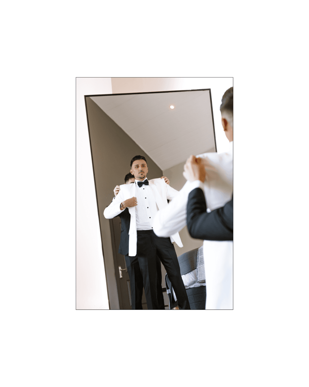 A groom in a white tuxedo jacket adjusts his bowtie while looking into a mirror