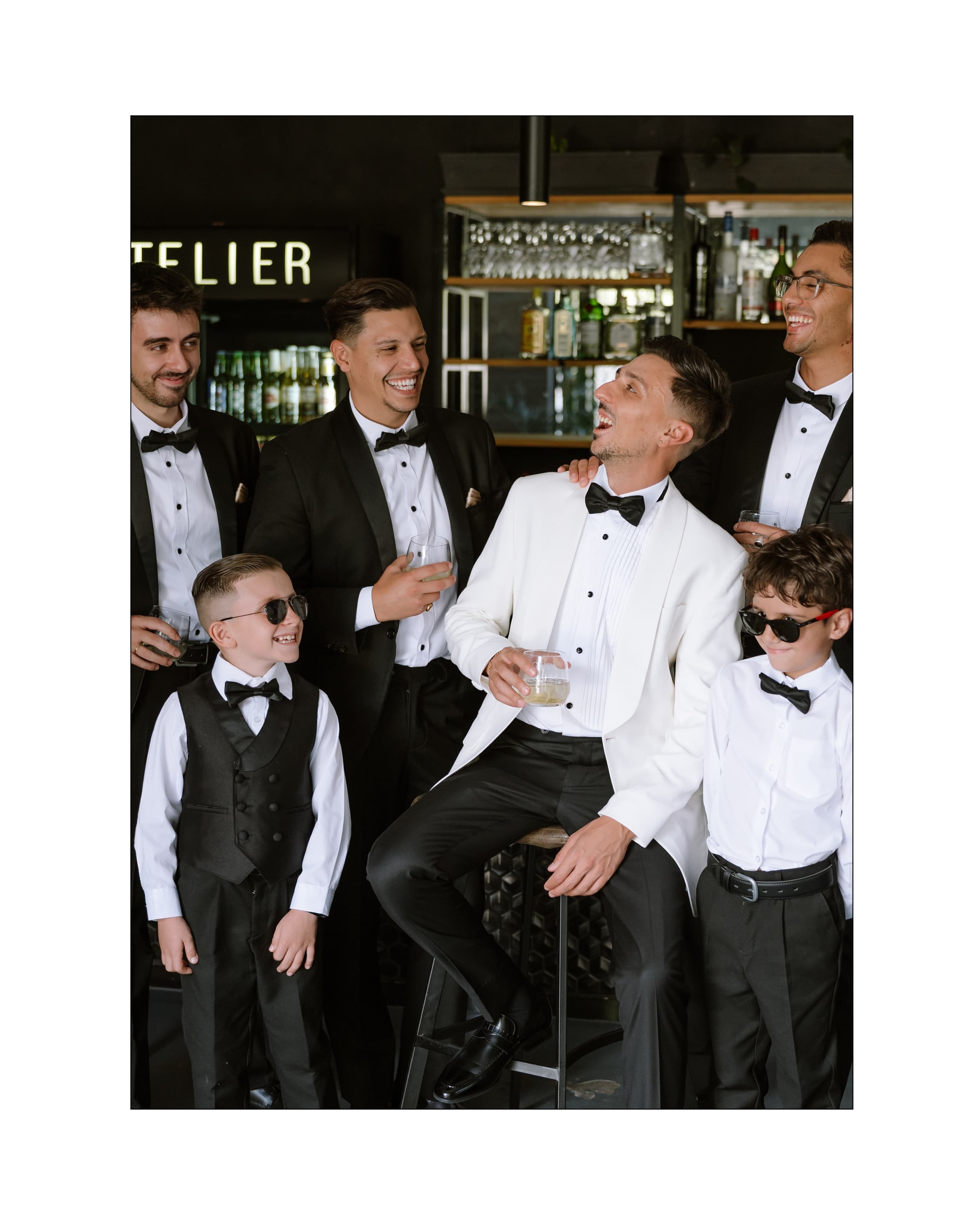 A groom in a white tuxedo laughs with his groomsmen and two boys at a bar