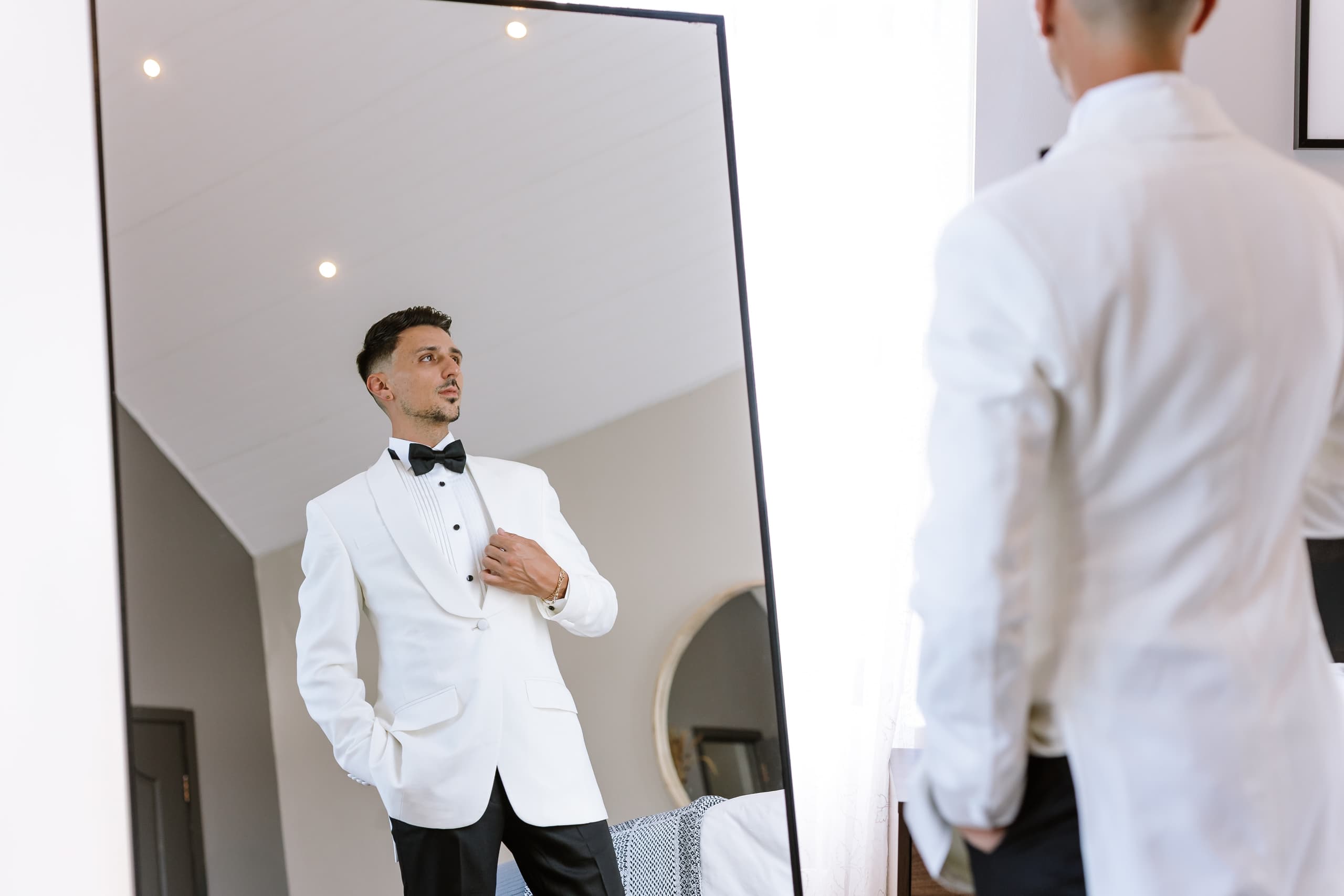 A groom in a white tuxedo jacket adjusts his lapel while looking into a mirror
