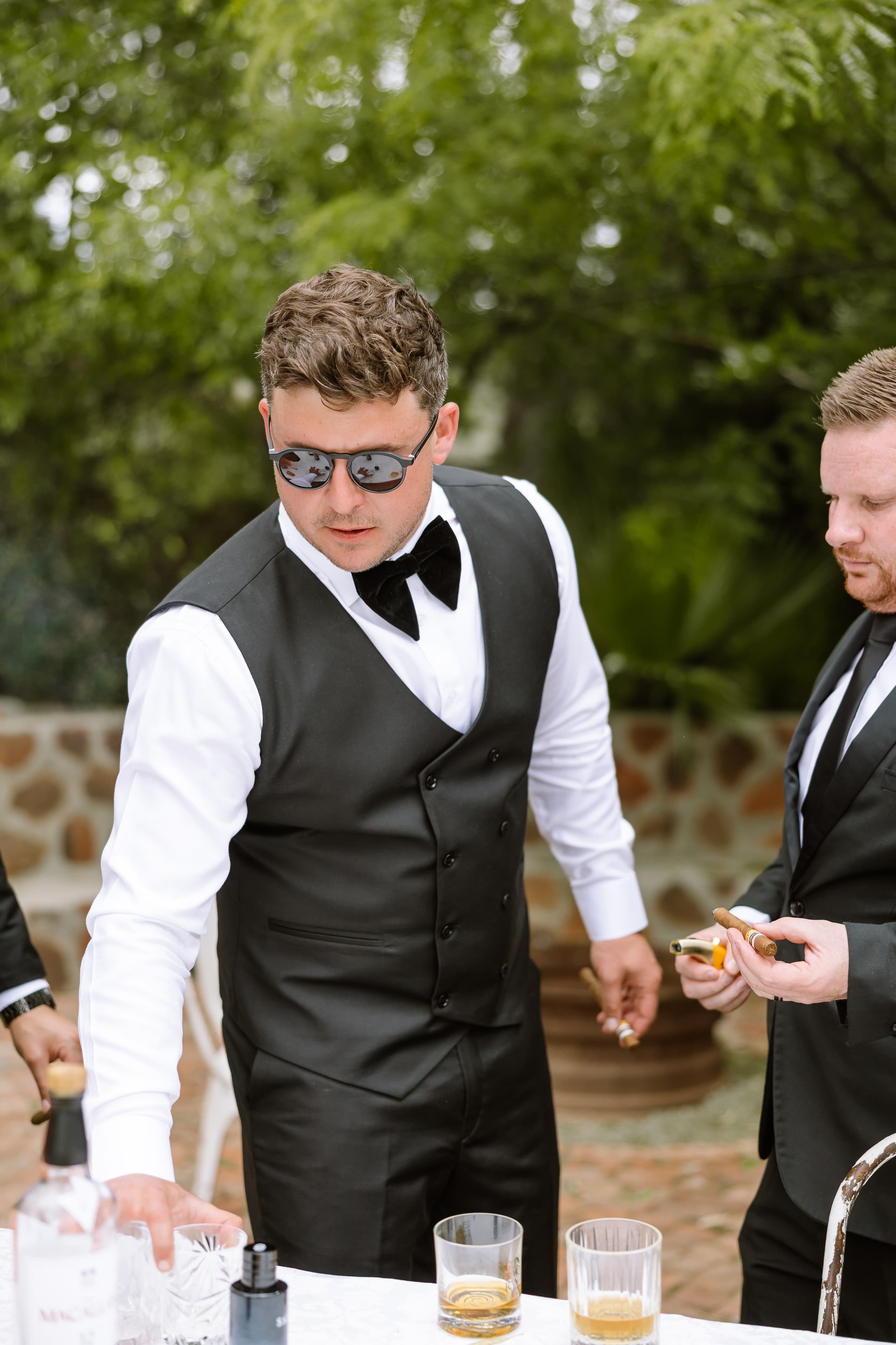 Groomsmen wearing formal black vests and bow ties prepare drinks and cigars outdoors