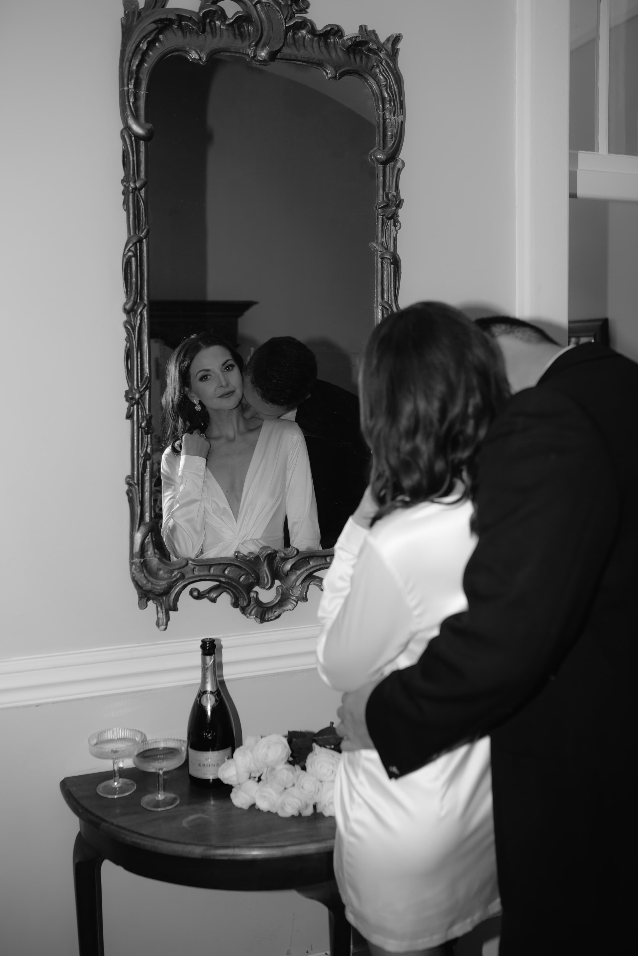 A bride in a silk robe looks into an ornate mirror while her partner kisses her neck