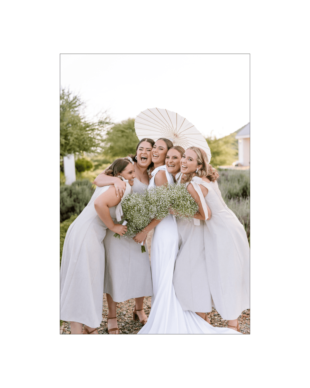 A joyful bride and her bridesmaids laughing together while holding bouquets under a white parasol
