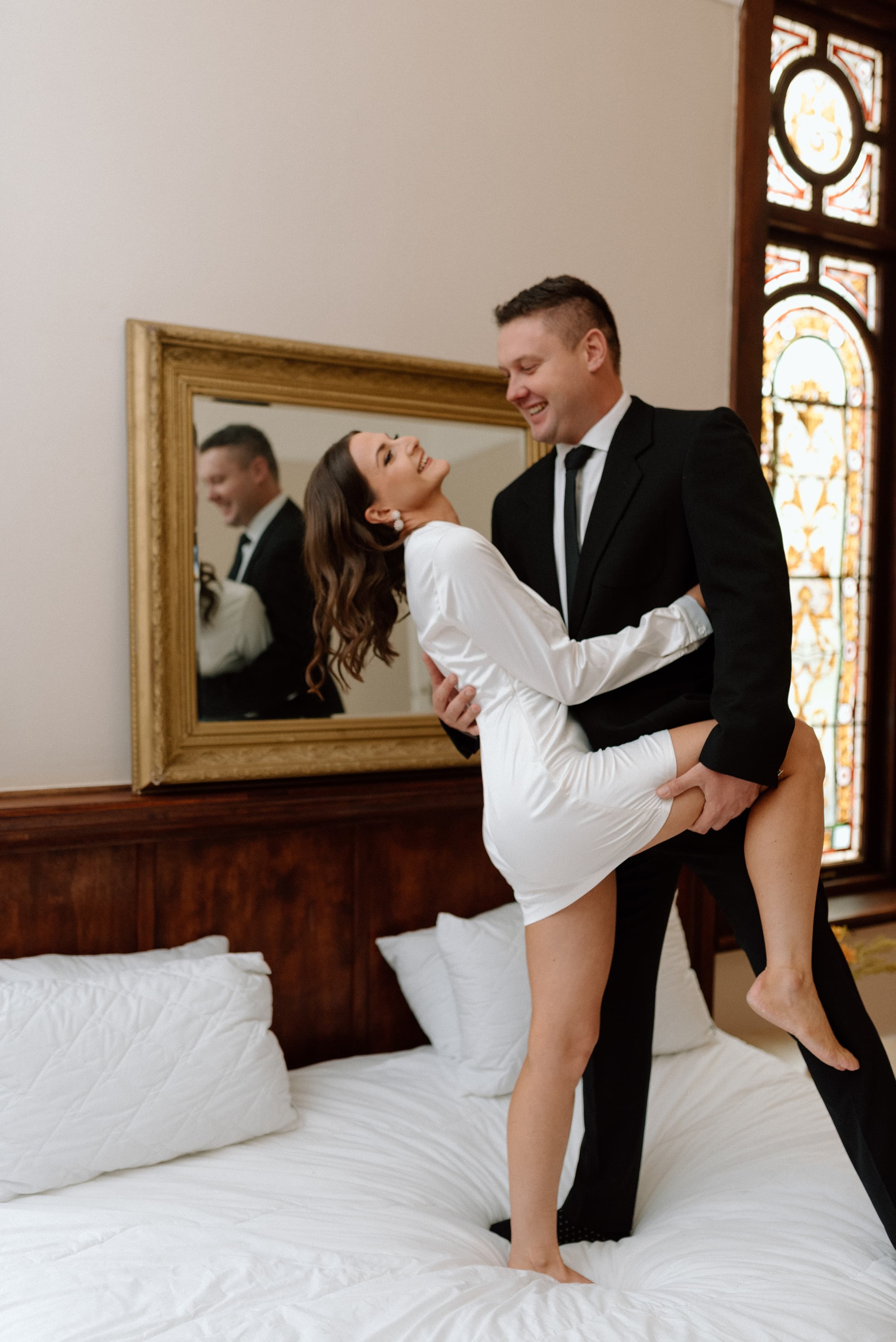 A joyful bride in a white dress dances with her groom in a bedroom