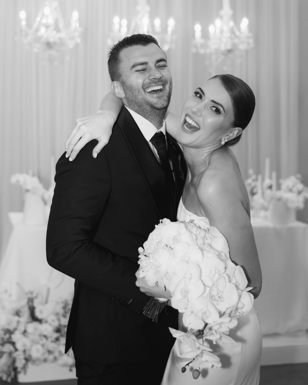 A joyful bride and groom laughing together while holding a large bouquet of white orchids
