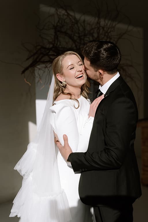 A laughing bride in a long-sleeved gown and veil, kissed on the cheek by her groom