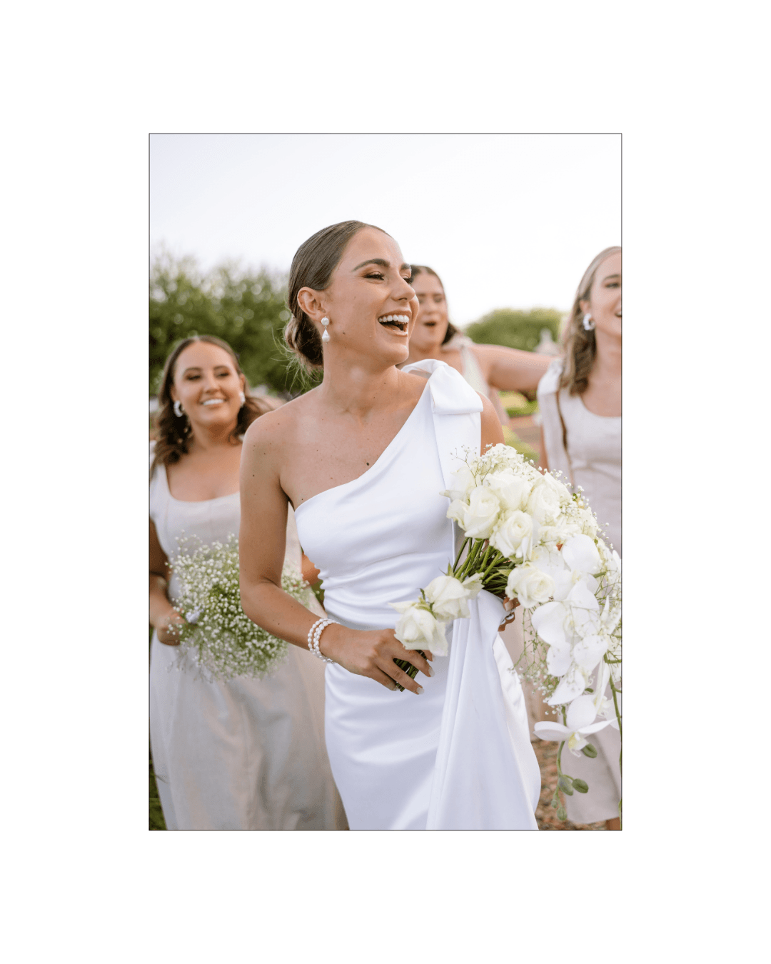 A joyful bride in a white one-shoulder gown laughs while walking with her bridesmaids