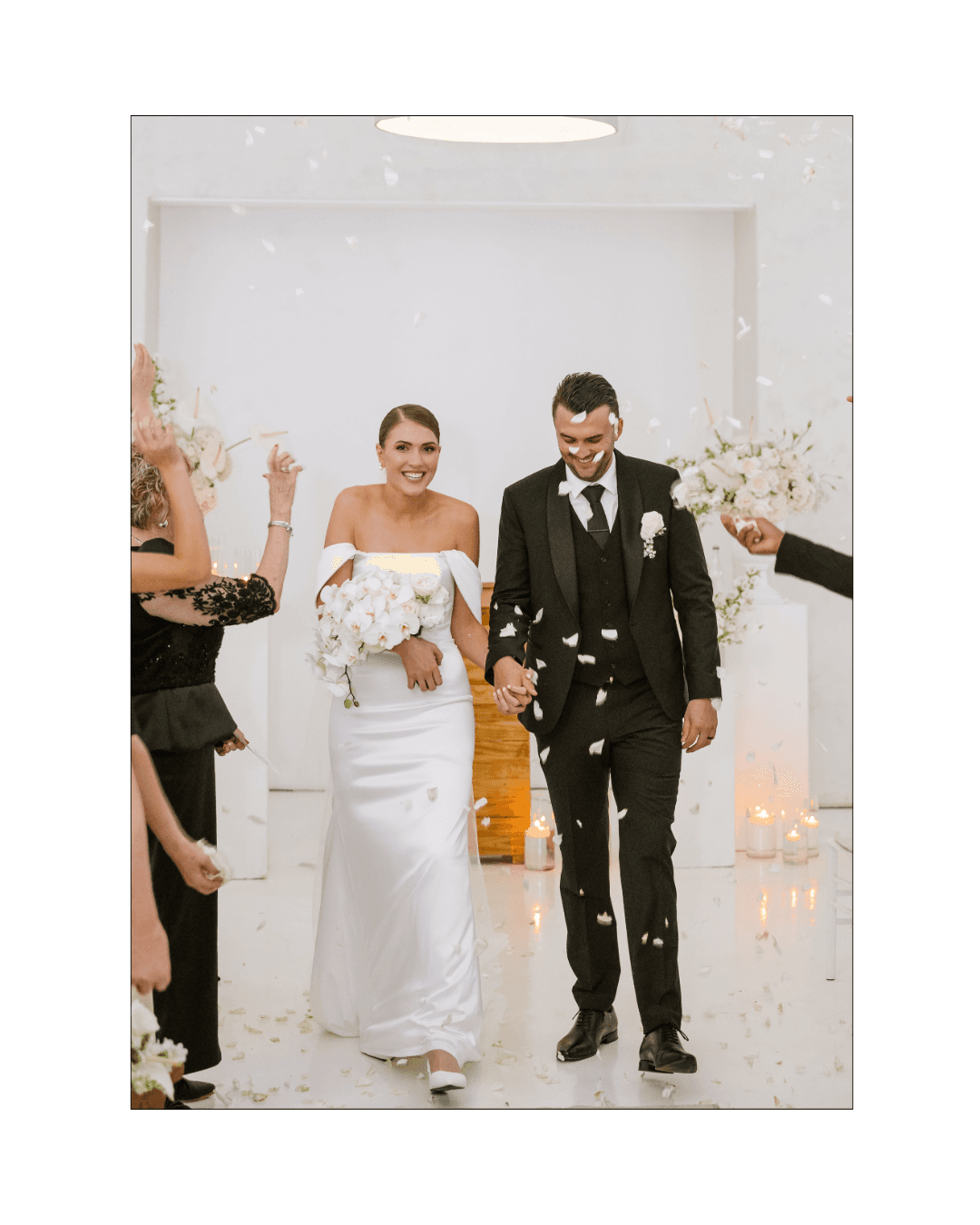 A joyful bride and groom walk hand in hand through a shower of white petals