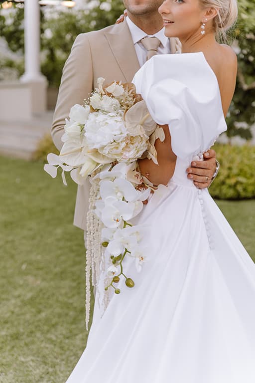 A bride holding a white orchid bouquet in her groom’s arms in a garden